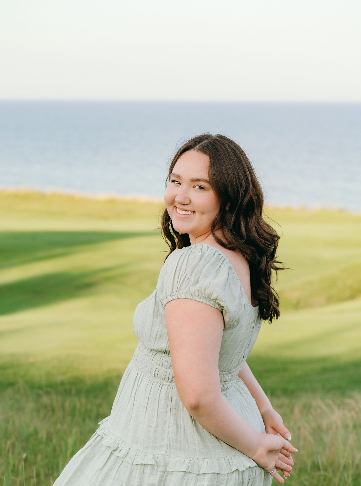 A young woman with long dark hair smiles while standing in a grassy field with the ocean in the background.