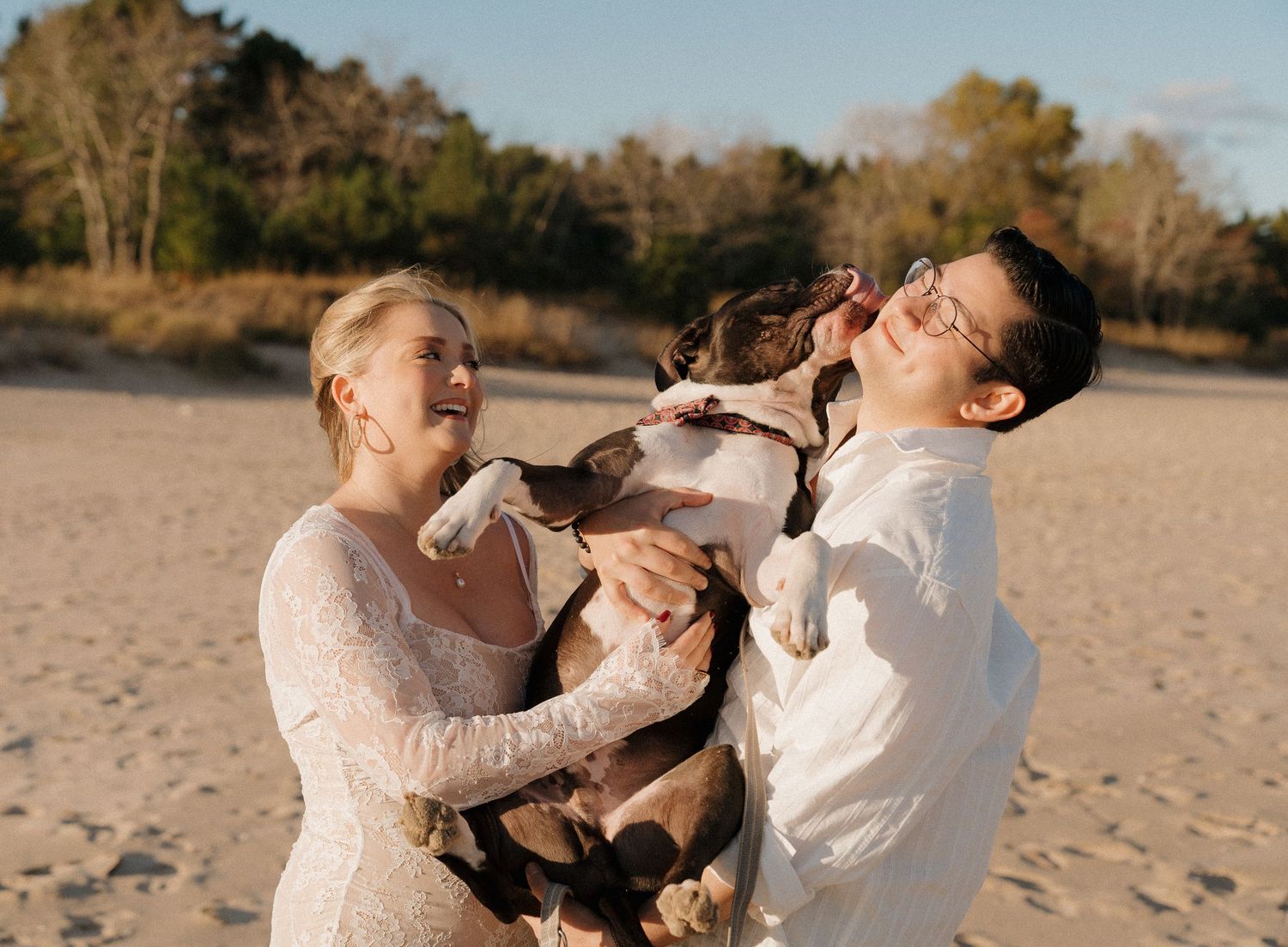 Joyful couple in white outfits laughing as dog licks their faces on a sandy beach.