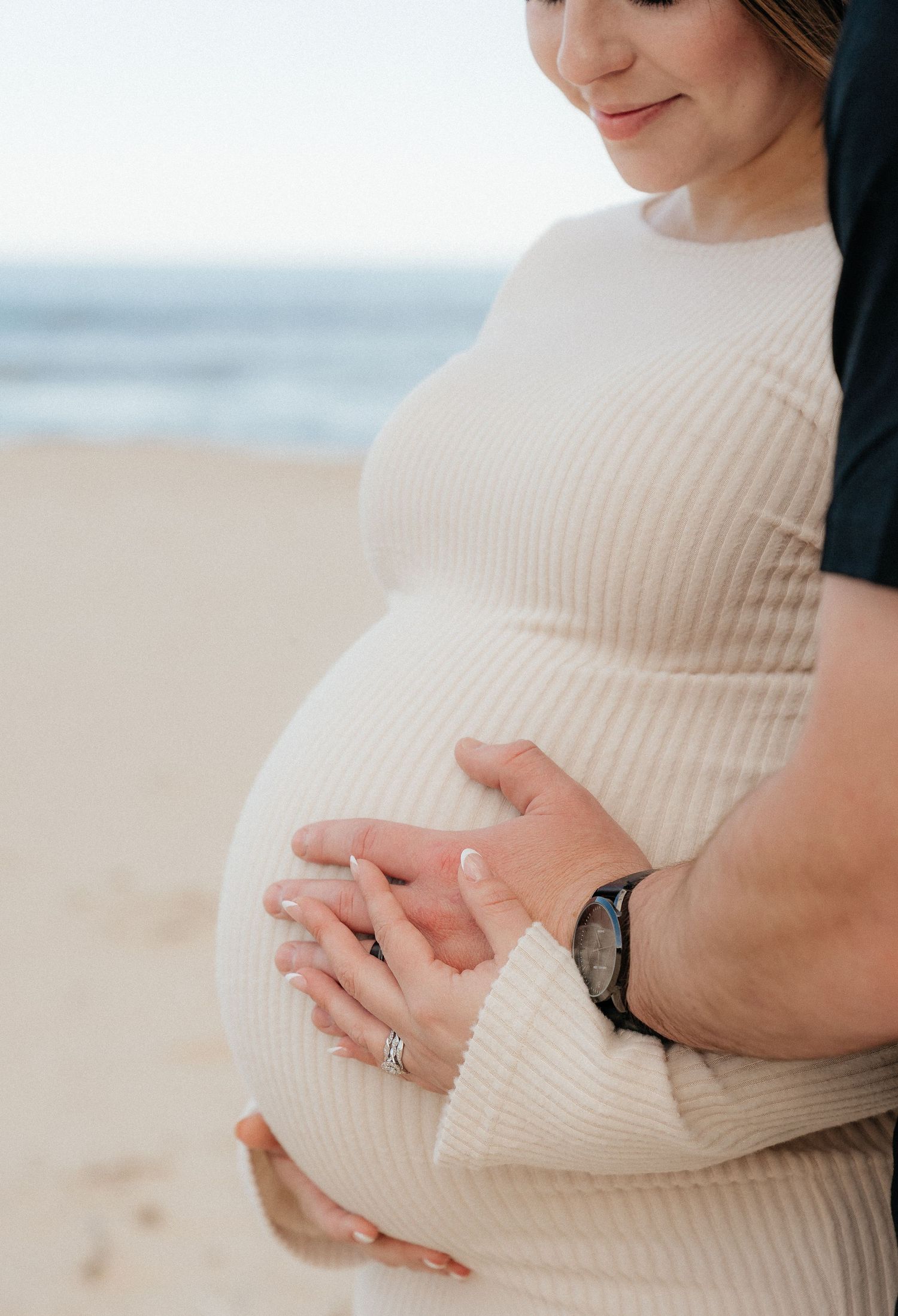 Pregnant woman in white ribbed dress cradling baby bump with partner on sandy beach.