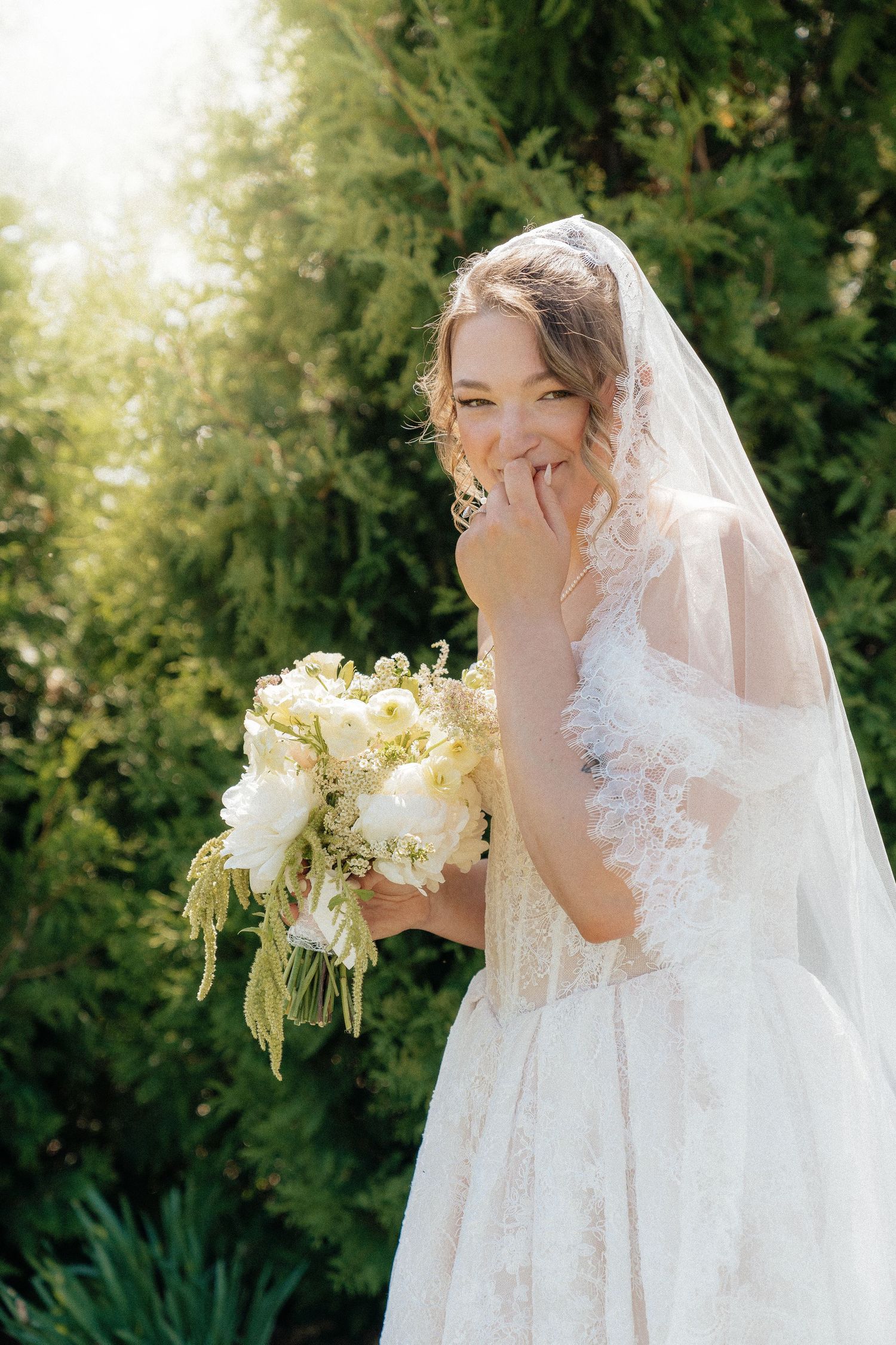 Laughing bride in lace wedding dress and veil holds white peony bouquet outdoors in sunlit garden.