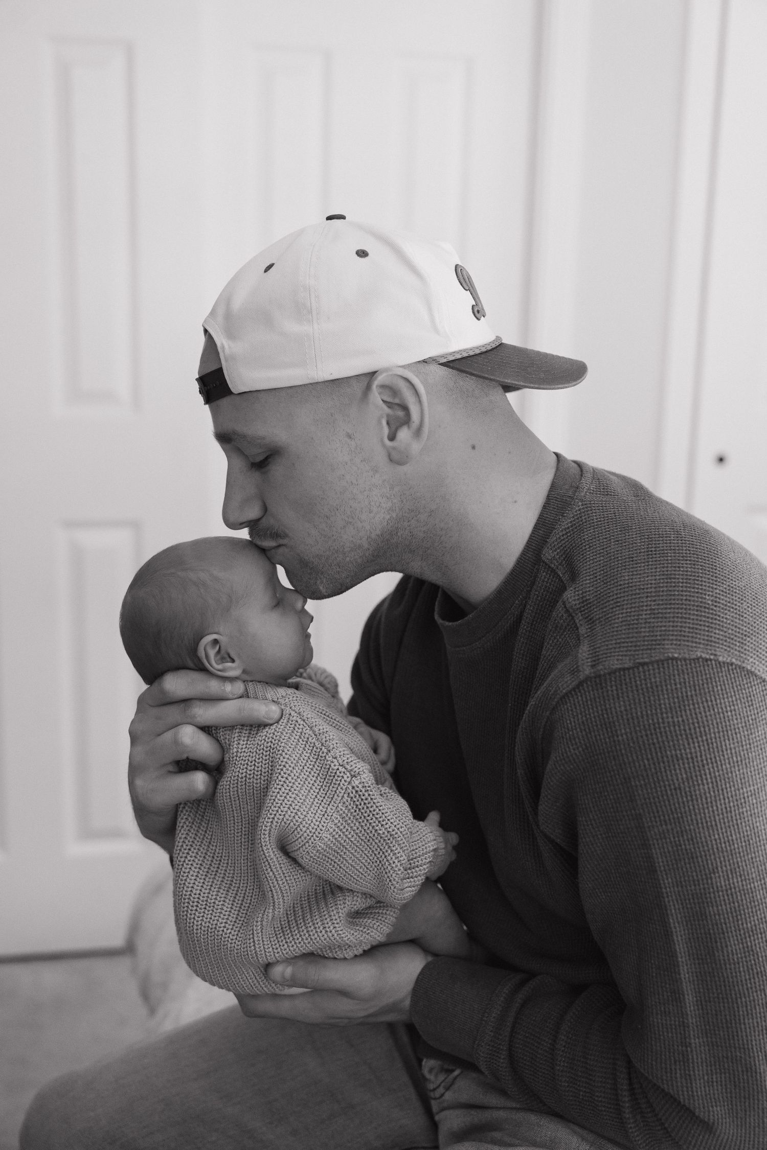 A man wearing a baseball cap is tenderly holding and gazing at a baby in a black and white photograph.