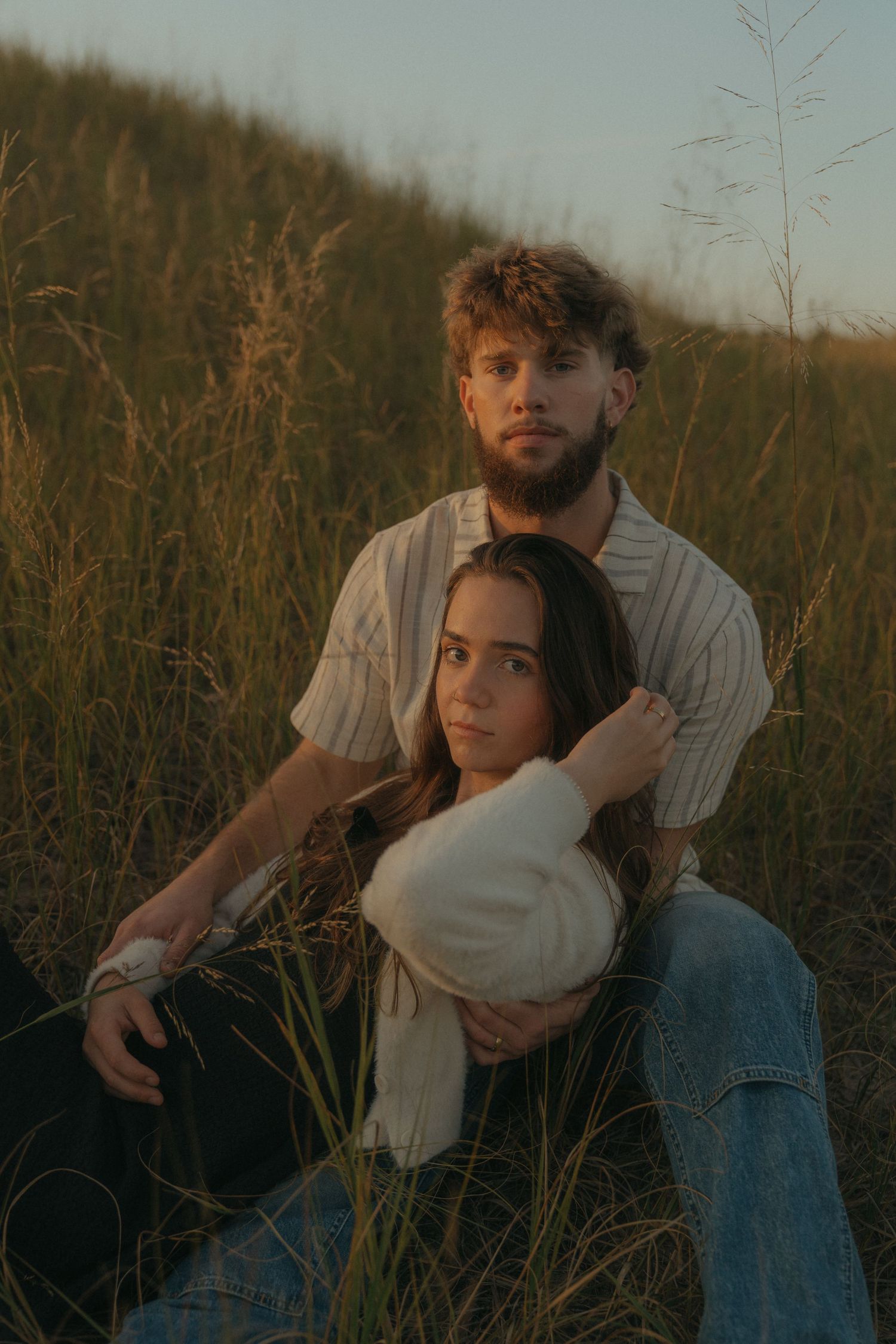 A man and woman sit together in a grassy field, embracing and enjoying the outdoors.