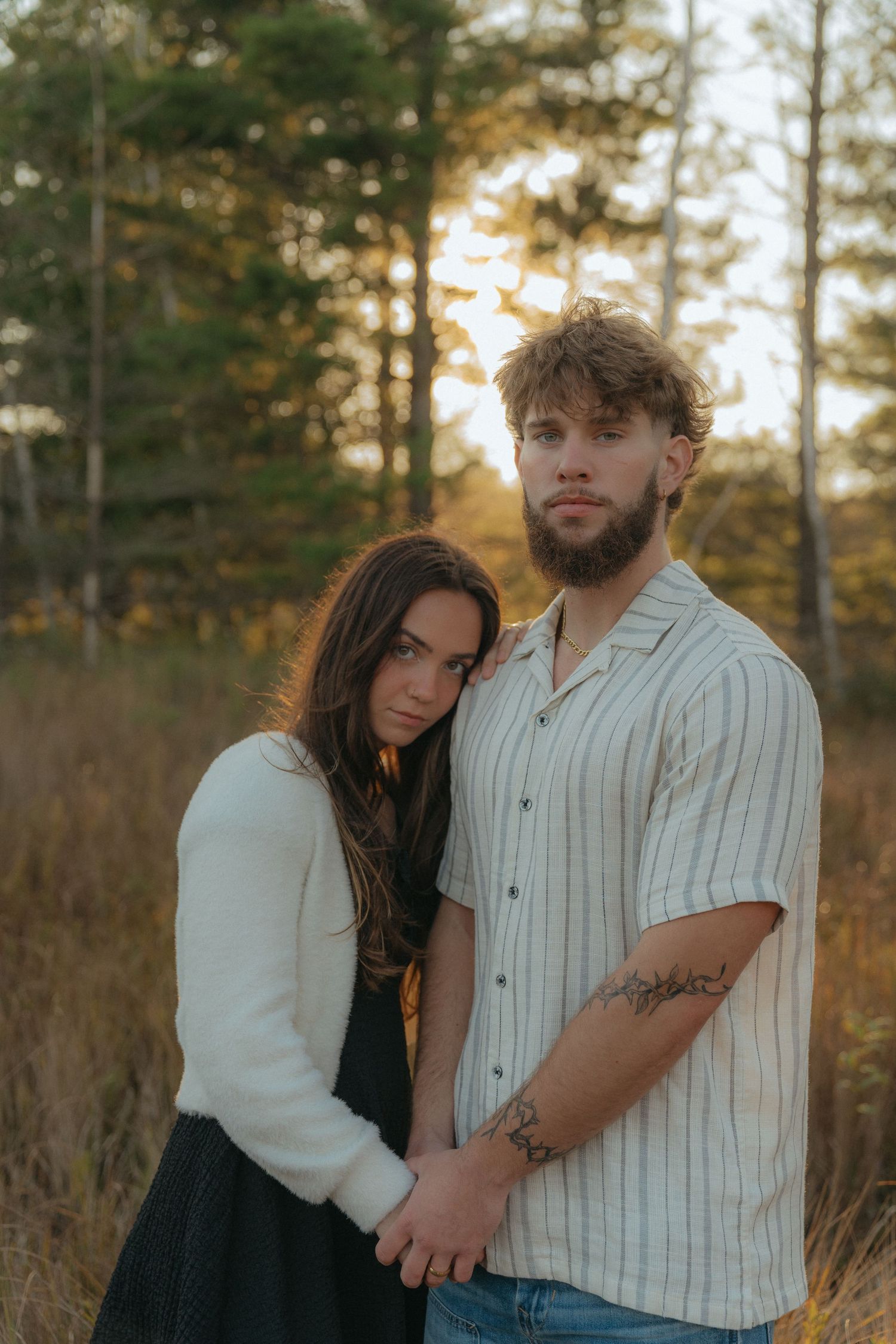 A young couple standing in a field surrounded by trees, the woman wearing a white sweater and the man wearing a striped shirt.