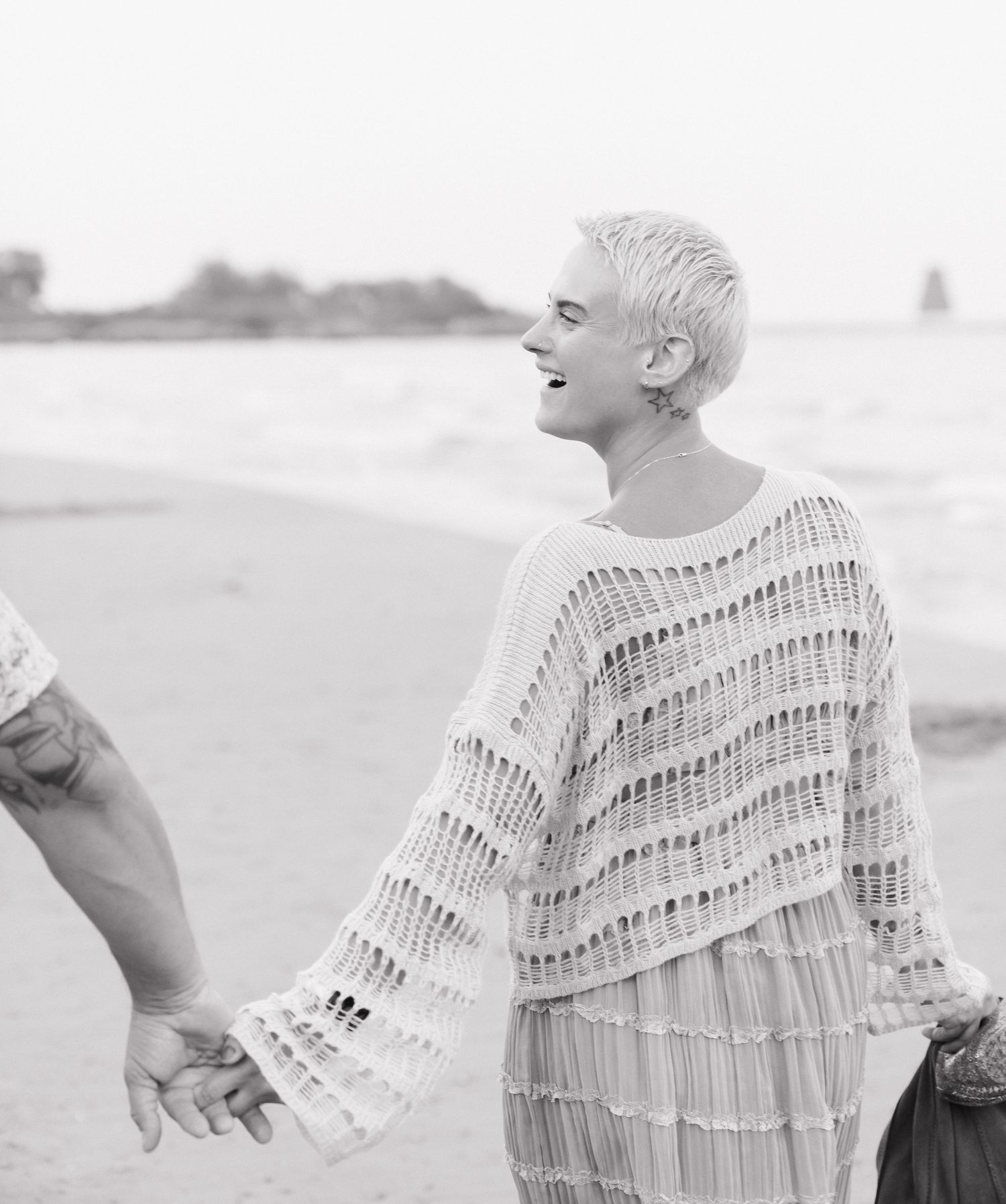 A person with short, blonde hair wearing a white, pleated outfit stands on a beach.