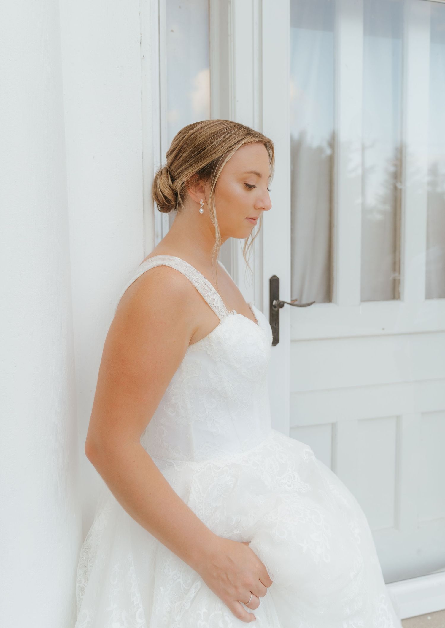 A woman wearing a white dress stands by a window, with her eyes closed.