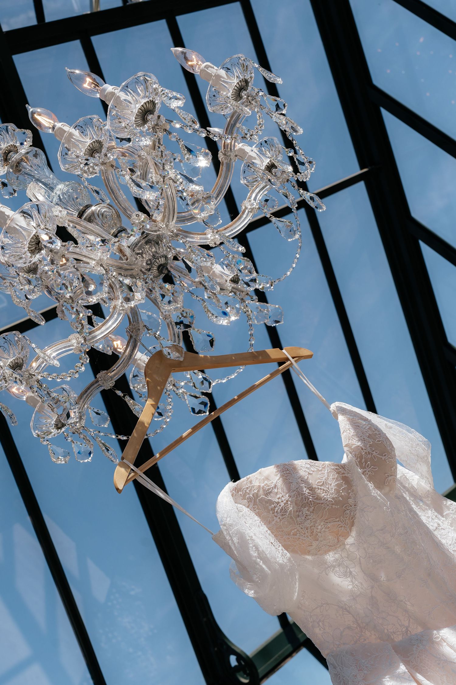 Crystal chandelier hanging from glass ceiling with elegant lace wedding dress on a wooden hanger below.