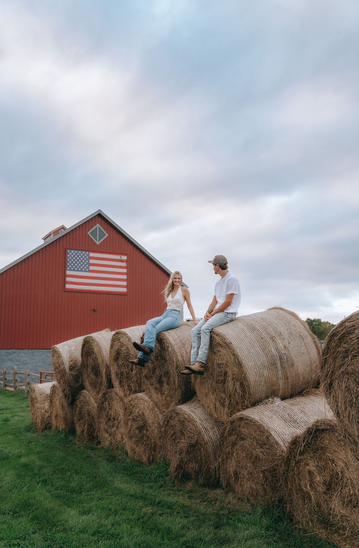 A couple sits on stacks of hay bales in a rural setting with a red barn and American flag in the background.