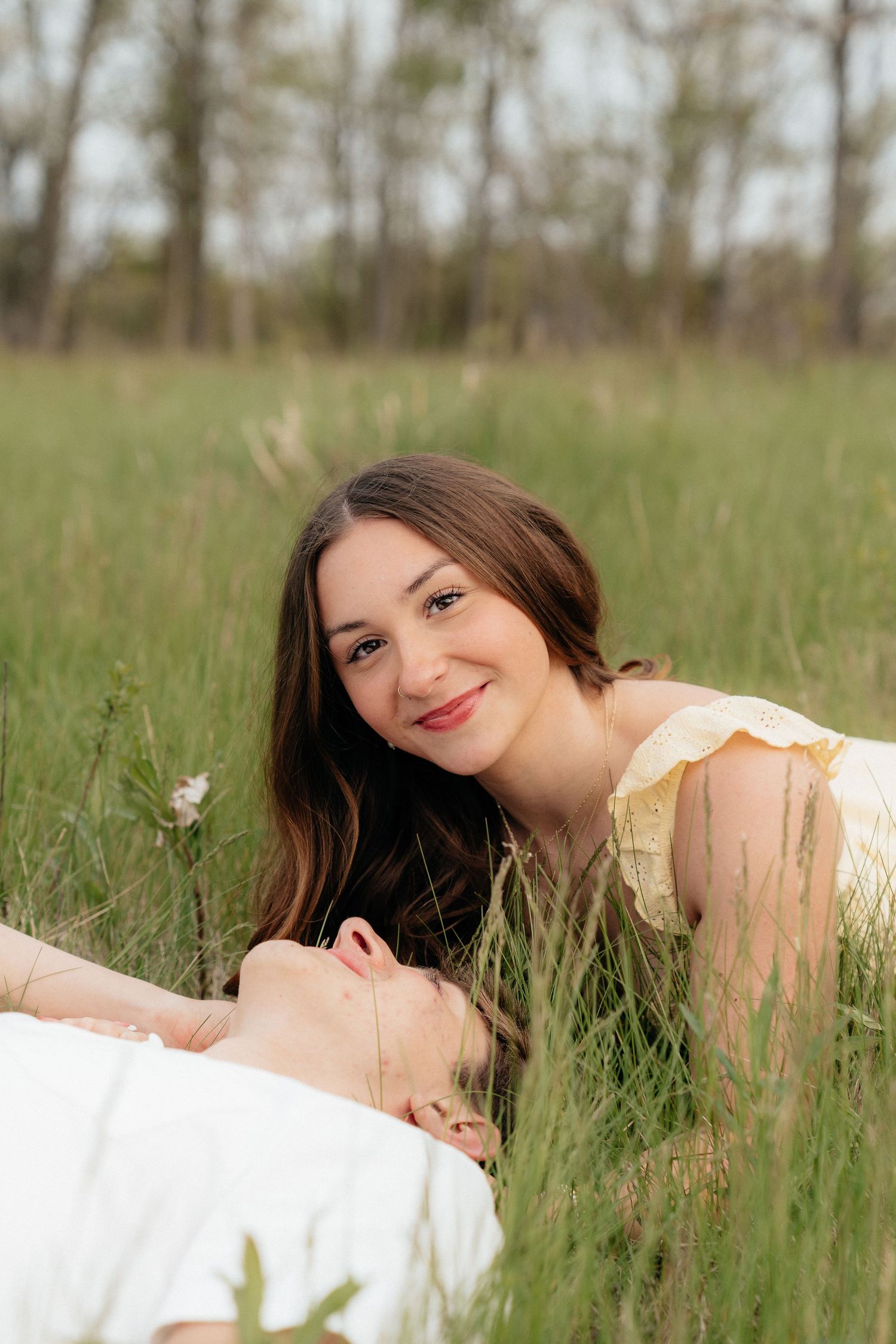 Two young women lying in tall green grass, one smiling at camera in yellow dress, trees in background.