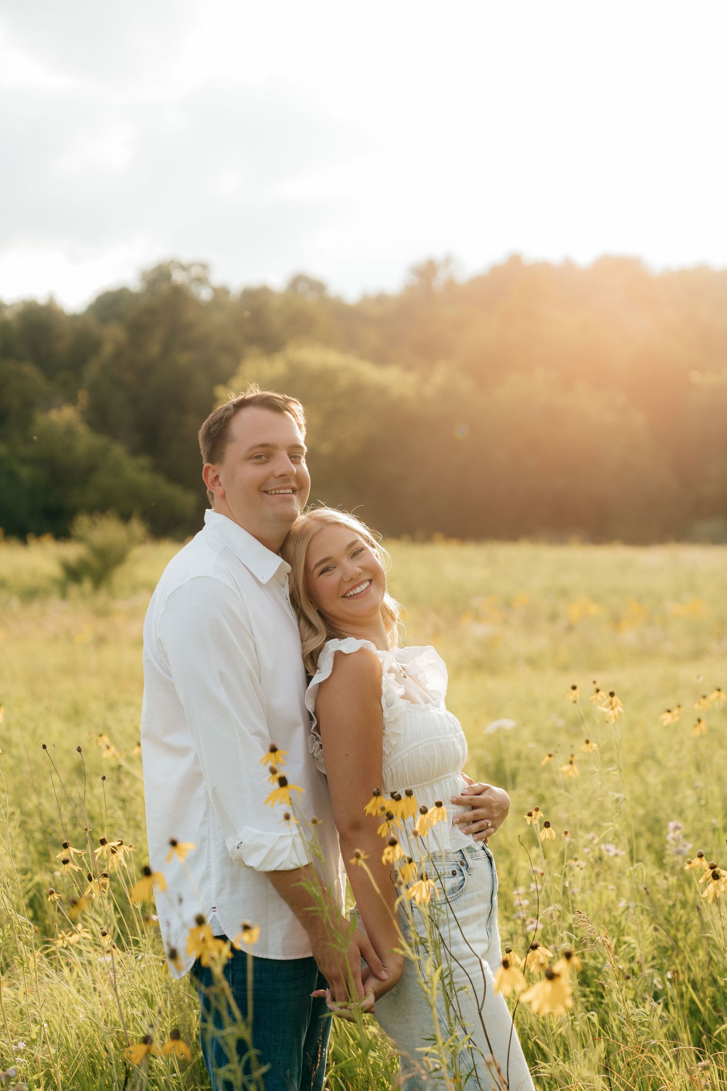 A smiling couple embraces in a sunlit wildflower field with yellow blooms, wearing white tops and jeans.