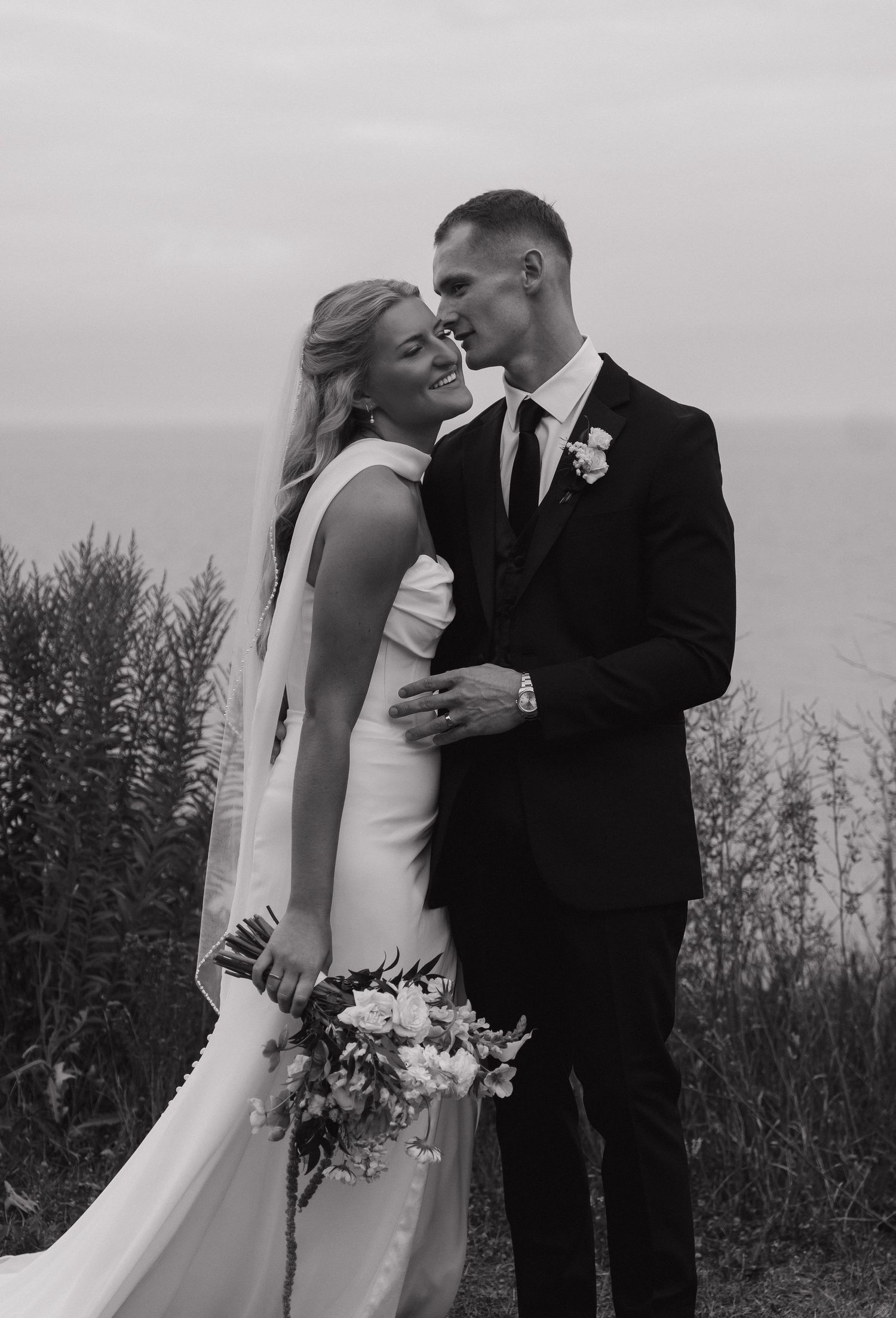 Black and white photo of a bride and groom sharing a romantic moment outdoors near the ocean on their wedding day.