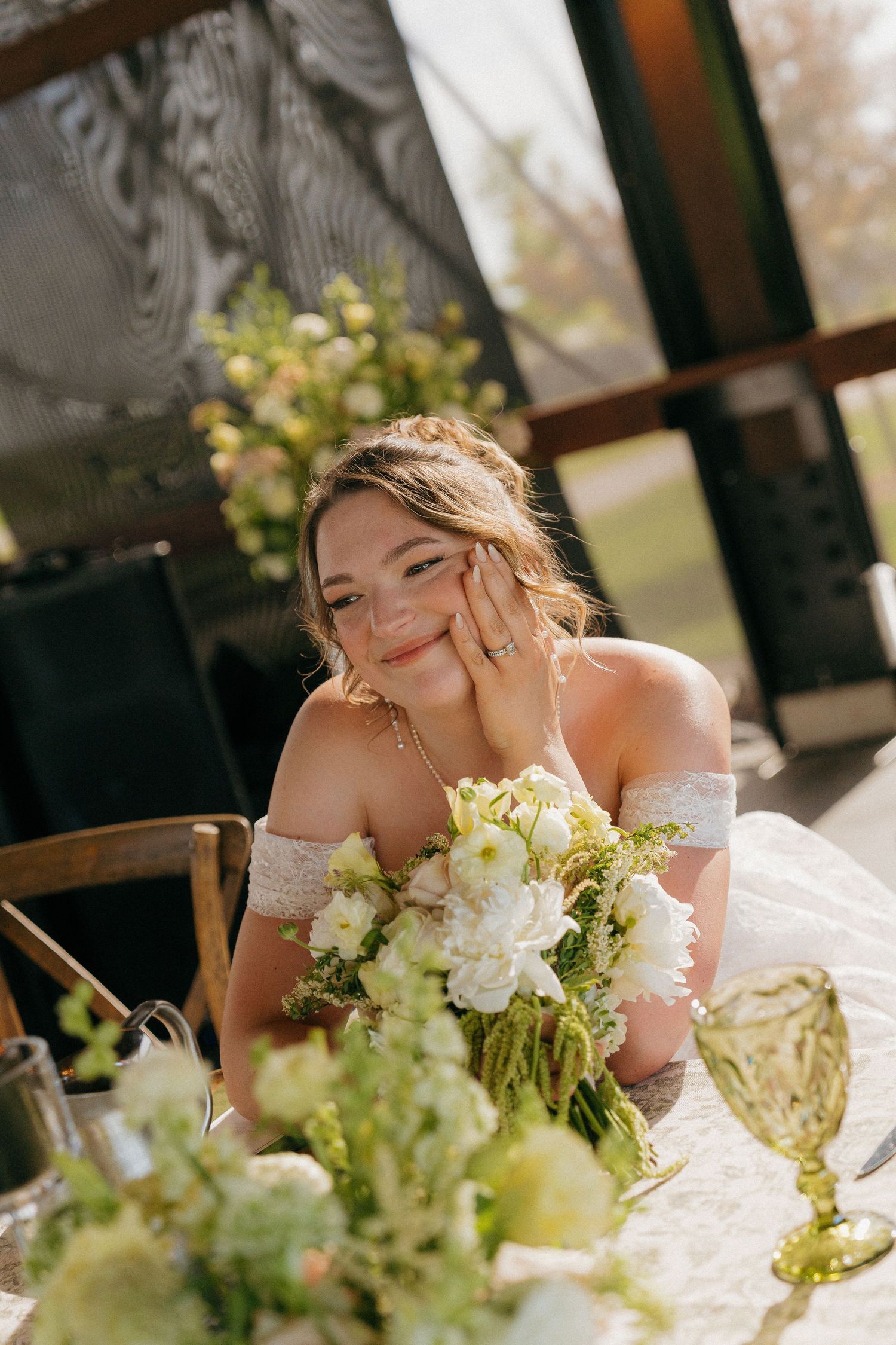 Smiling bride in off-shoulder lace gown holds white floral bouquet while seated at elegant reception table.