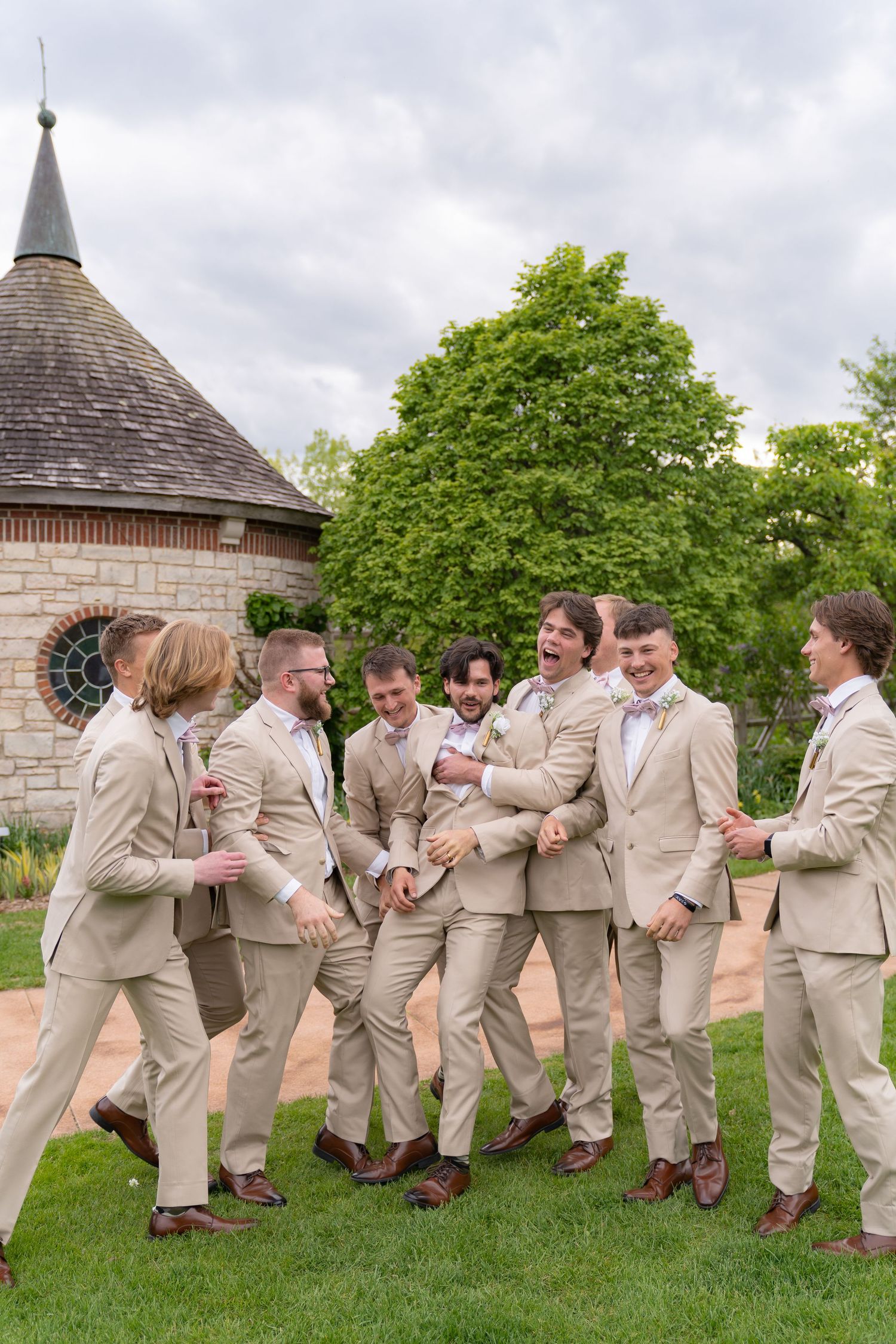 A group of men in tan suits stand together in a grassy outdoor setting with a tiled roof structure in the background.