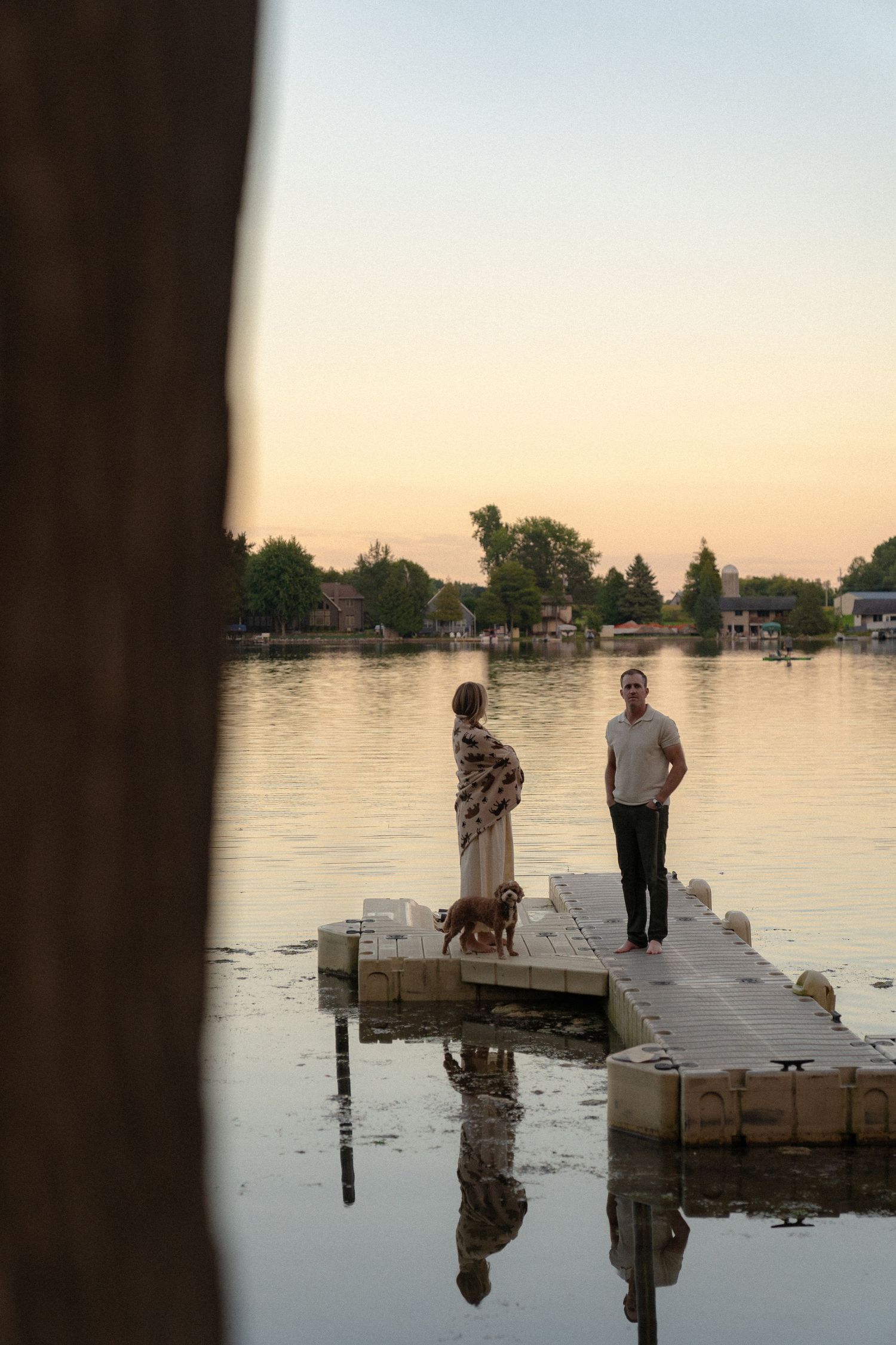 A couple standing on a wooden dock, reflected in the still water at sunset.