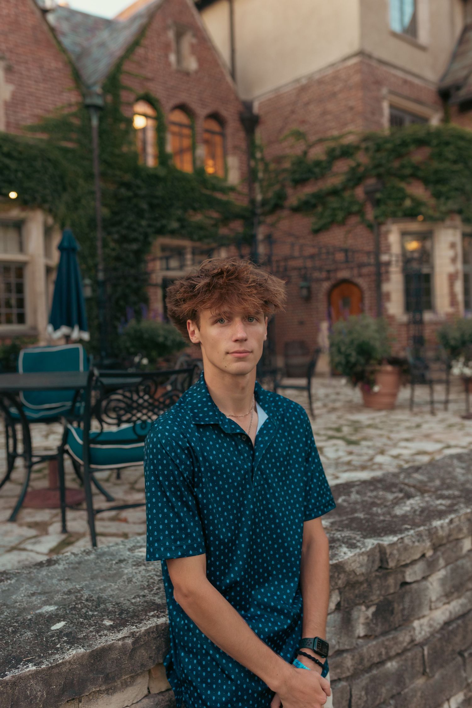 A young man with curly hair stands on a cobblestone street in front of a brick building with arched windows.