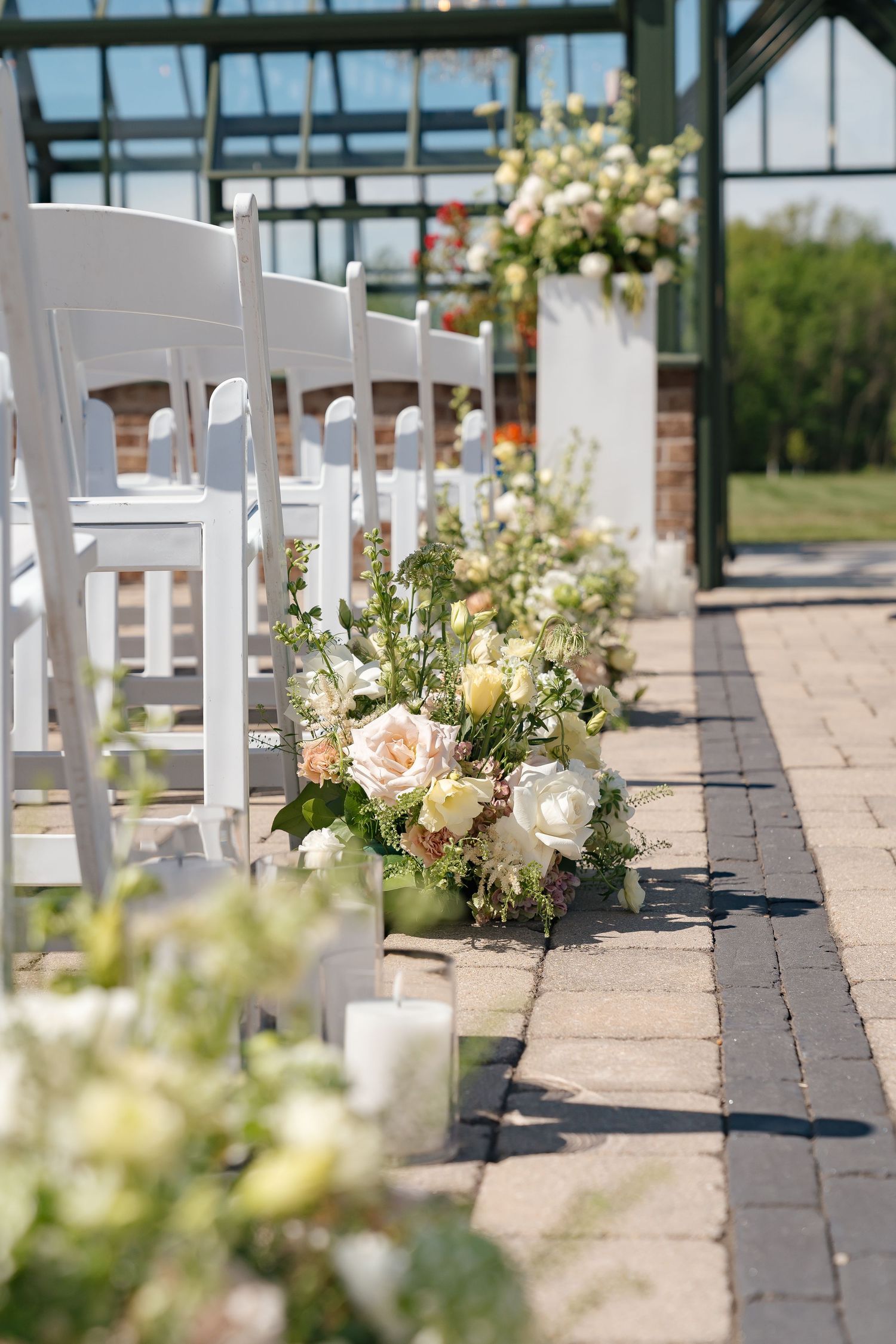 White chairs line a brick aisle adorned with lush floral arrangements and candles at an elegant outdoor wedding ceremony.