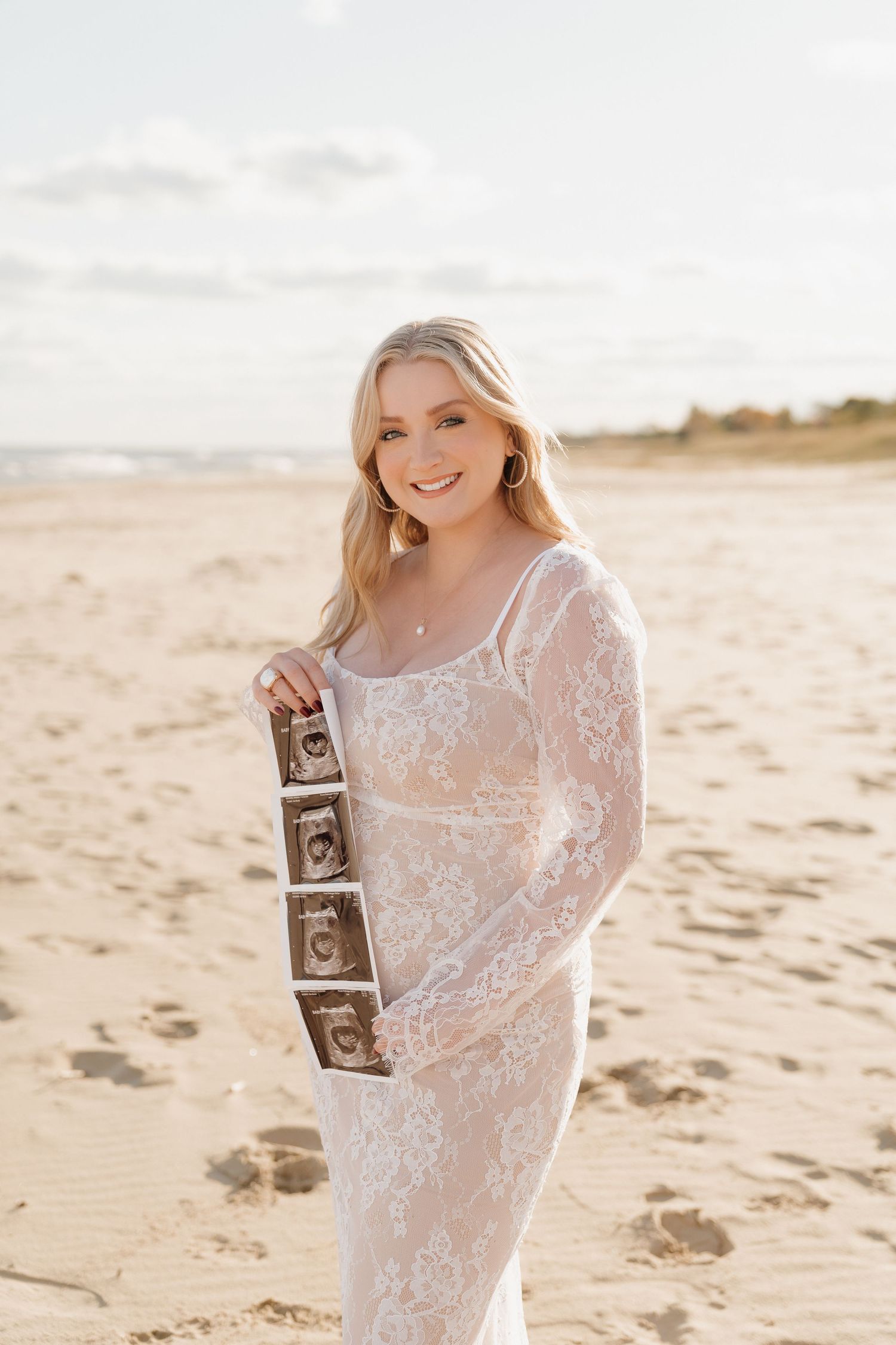 Pregnant woman in white lace dress holding ultrasound photos on a sandy beach at golden hour.