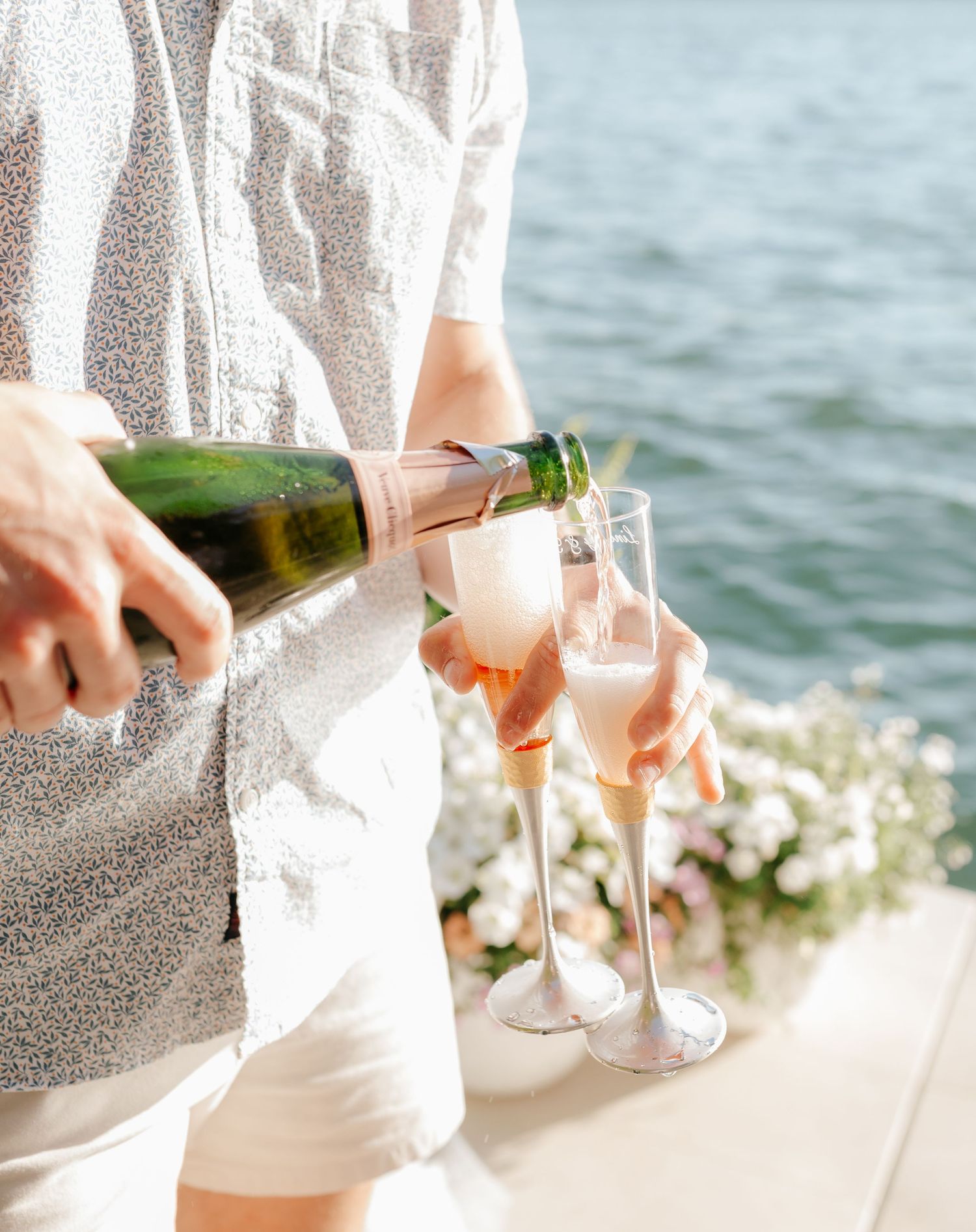 A person is pouring champagne into glasses while standing near the water's edge on a sunny day.