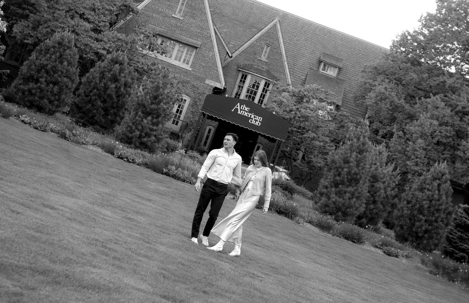 A black and white photograph shows a couple walking on a grassy path in front of a building.