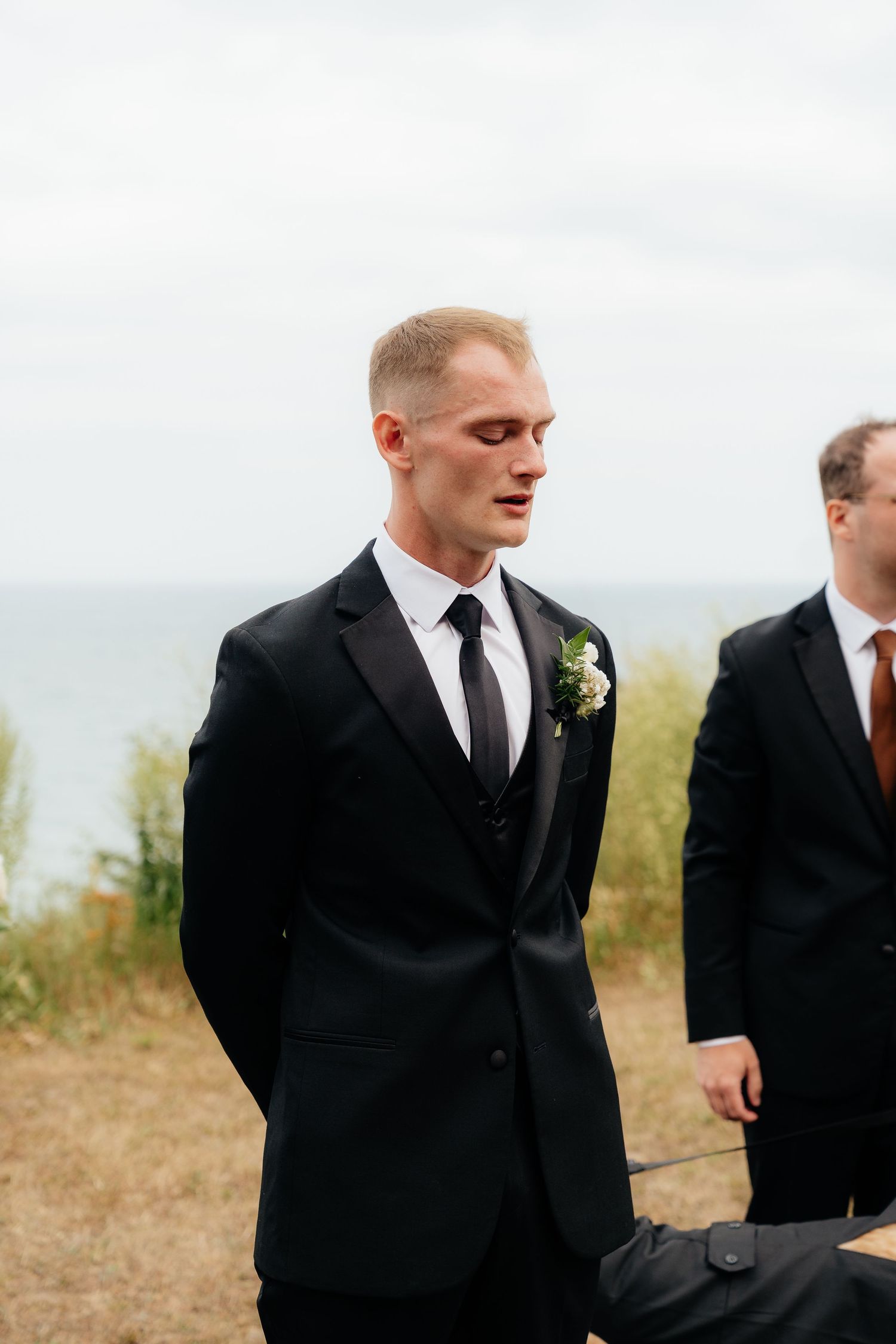 Groom in black tuxedo with floral boutonniere stands outdoors near ocean, with groomsman visible in background.