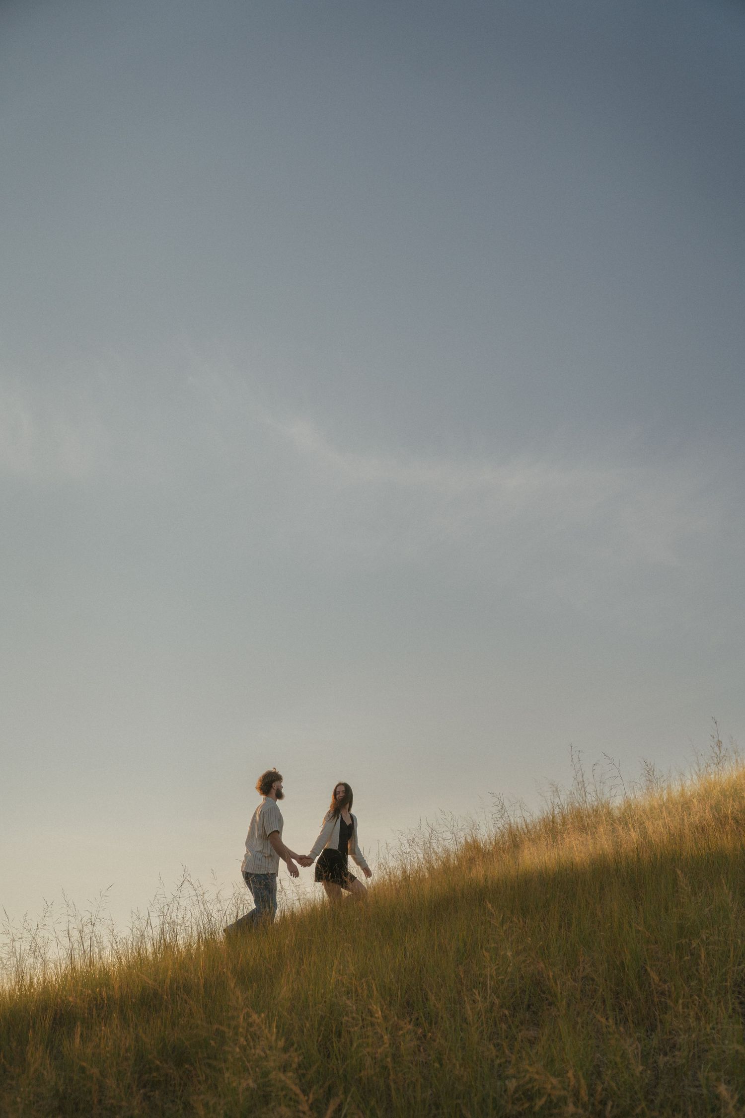 Two people walking hand-in-hand on a grassy hill against a cloudy sky.