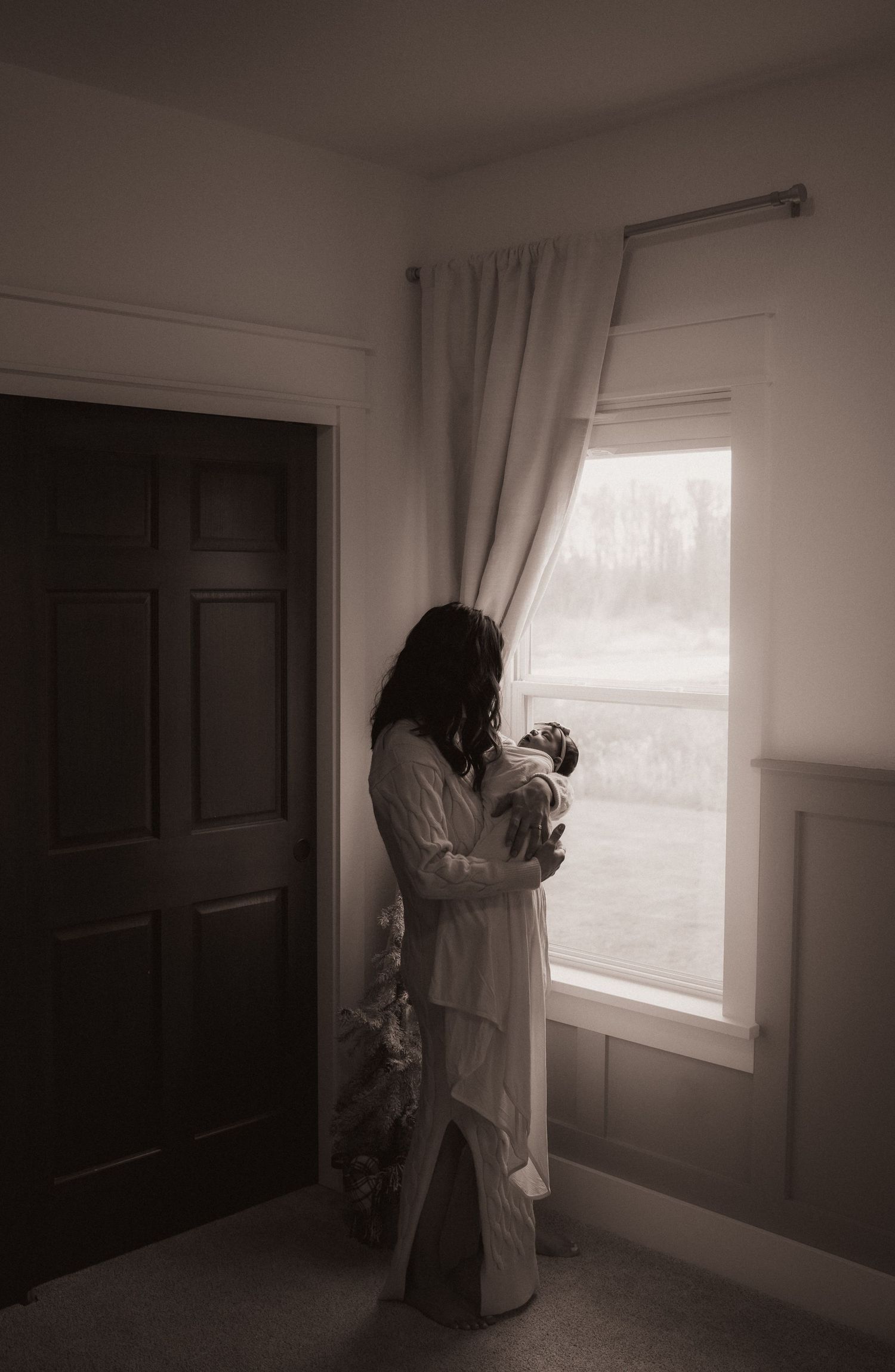 A woman in a white dress stands near a window, holding a baby in her hands, in a dimly lit room.