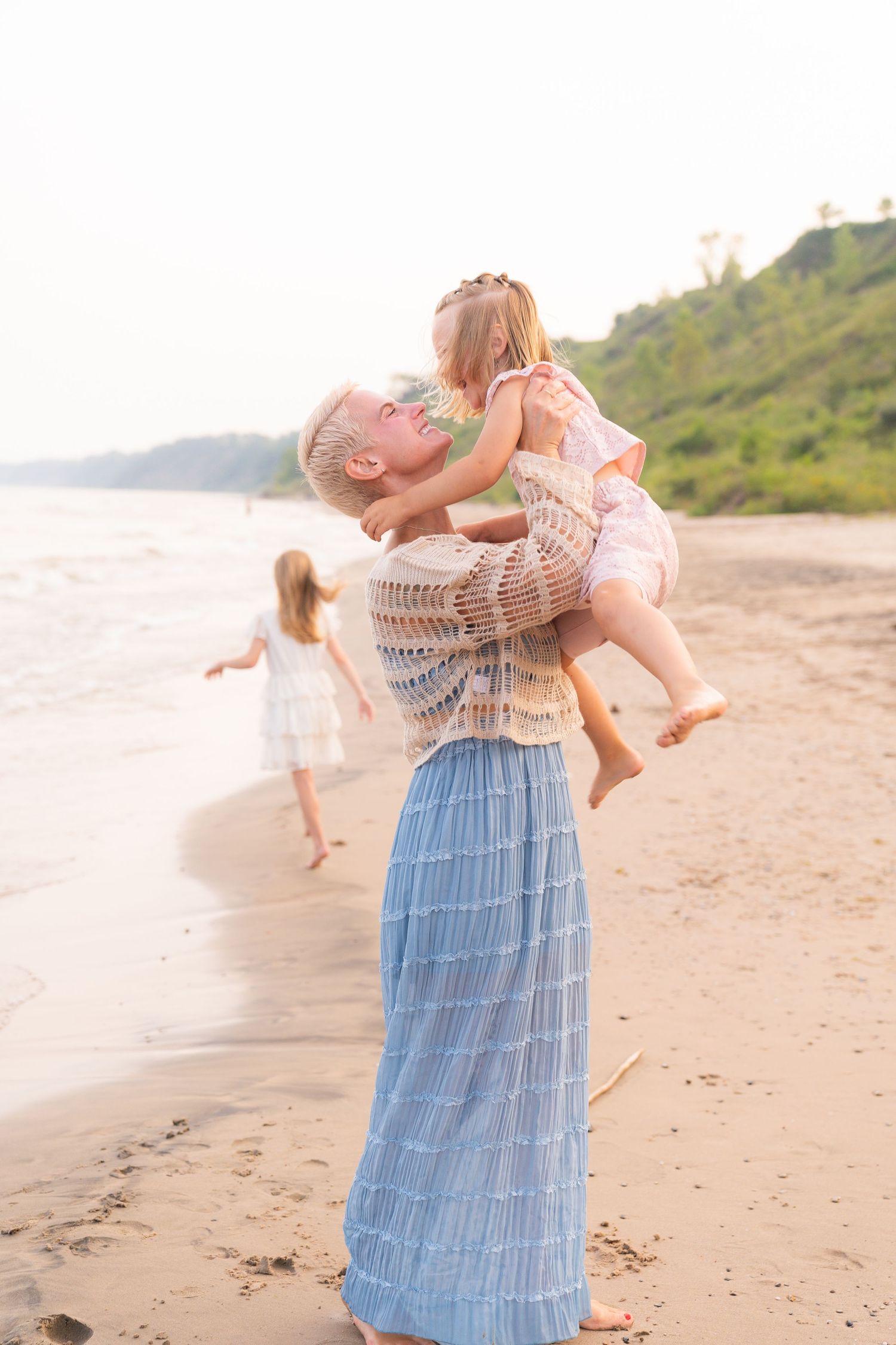 A woman lifts a young girl in the air on a beach, with another child visible in the background.