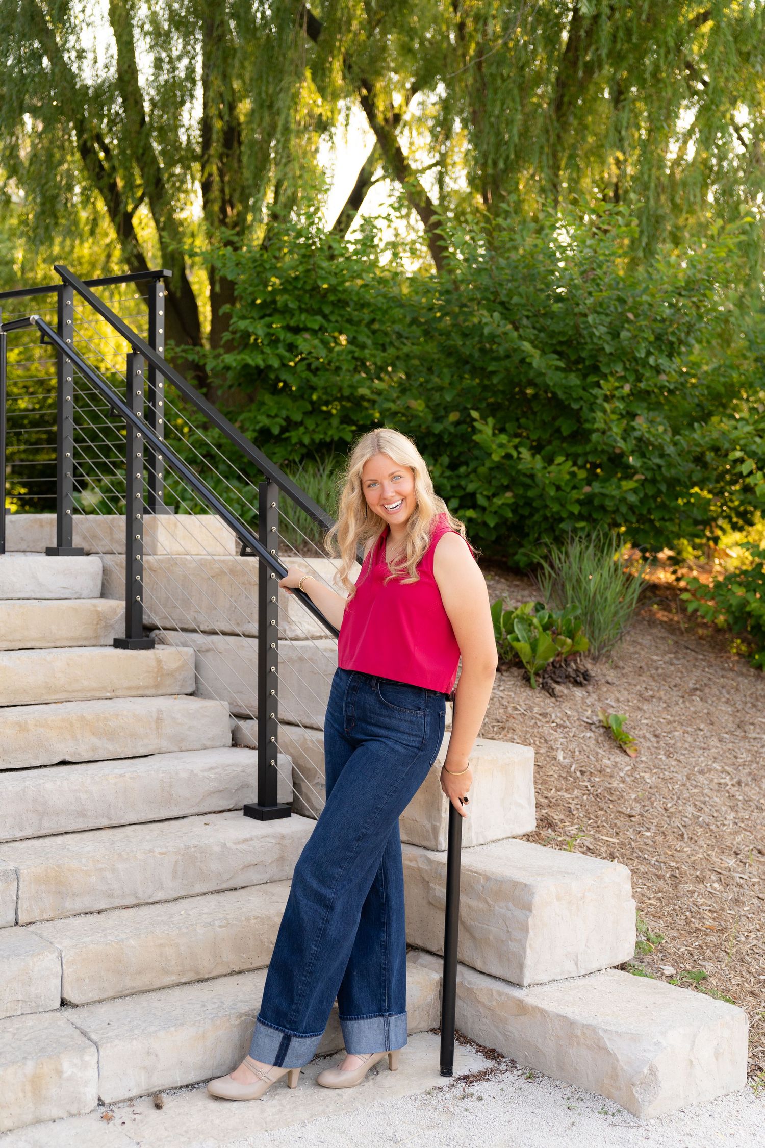 A smiling woman wearing a red shirt and jeans stands on outdoor stairs surrounded by lush green trees.