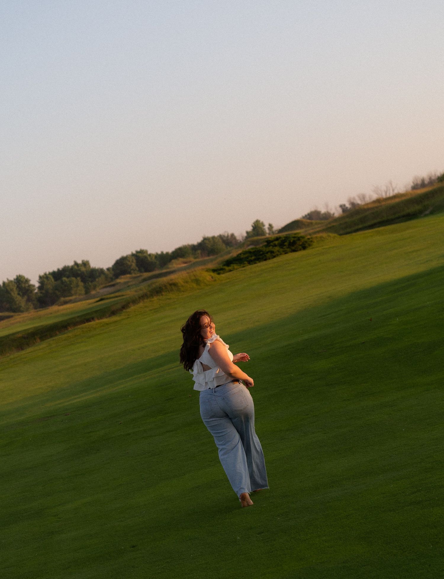 A woman walks through a grassy field with a scenic landscape in the background.