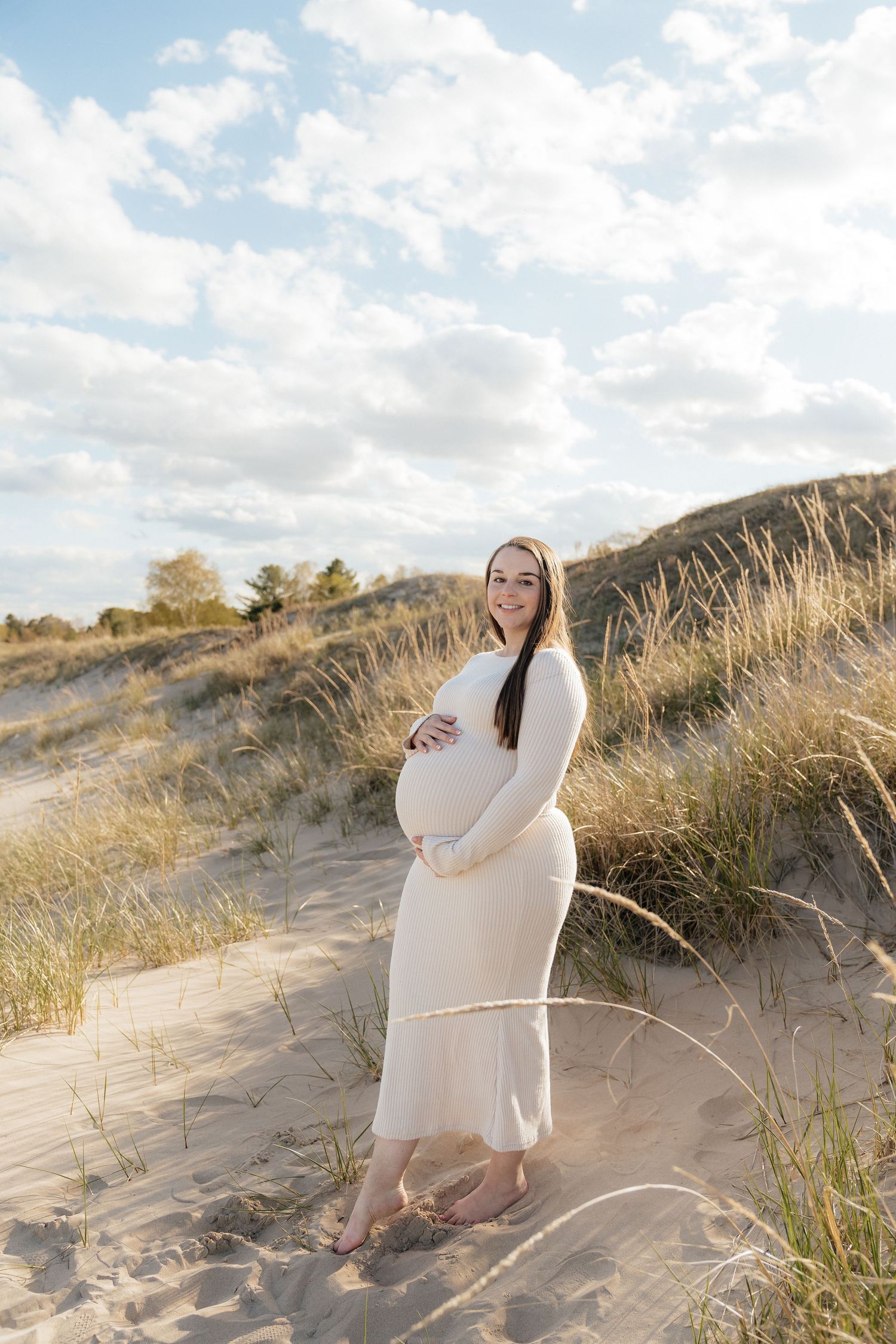 Pregnant woman in white dress cradling baby bump on sandy beach dunes with grass and cloudy sky.