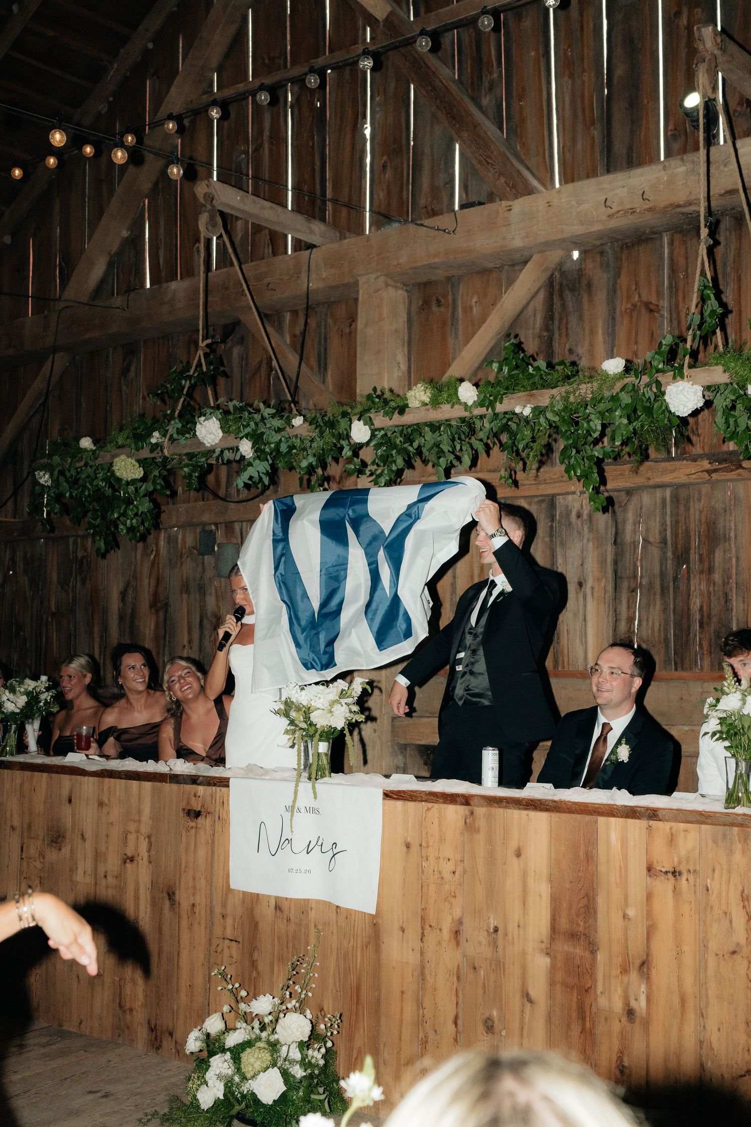 Couple holding NY flag at rustic barn wedding reception with greenery garland and white floral arrangements.