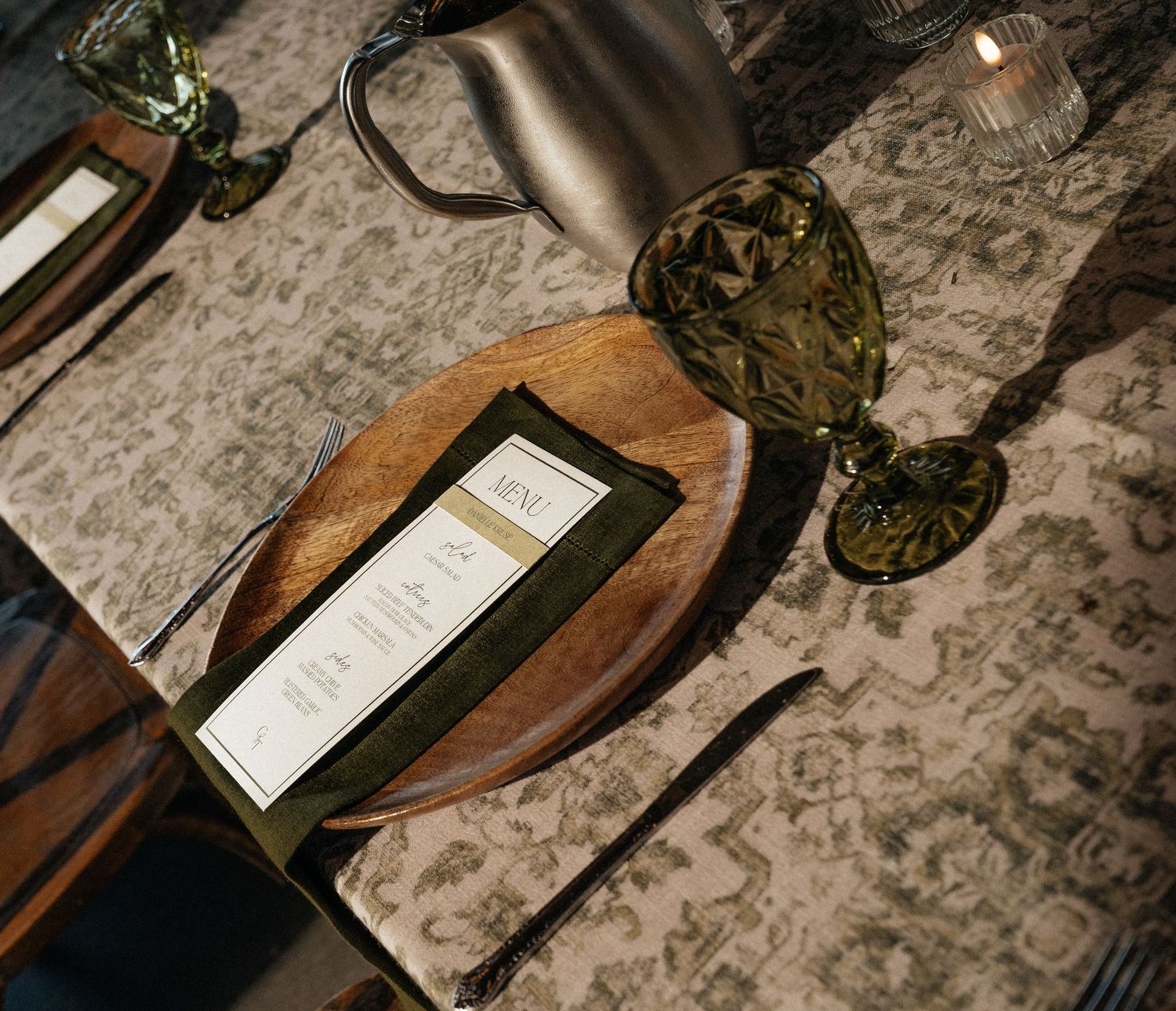 Elegant table setting with wooden charger plate, olive green napkin, menu card, and dark green crystal goblet.