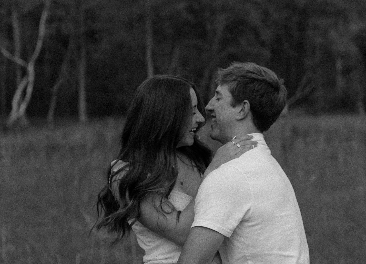 Black and white photo of a romantic couple embracing and smiling at each other in an outdoor field setting.
