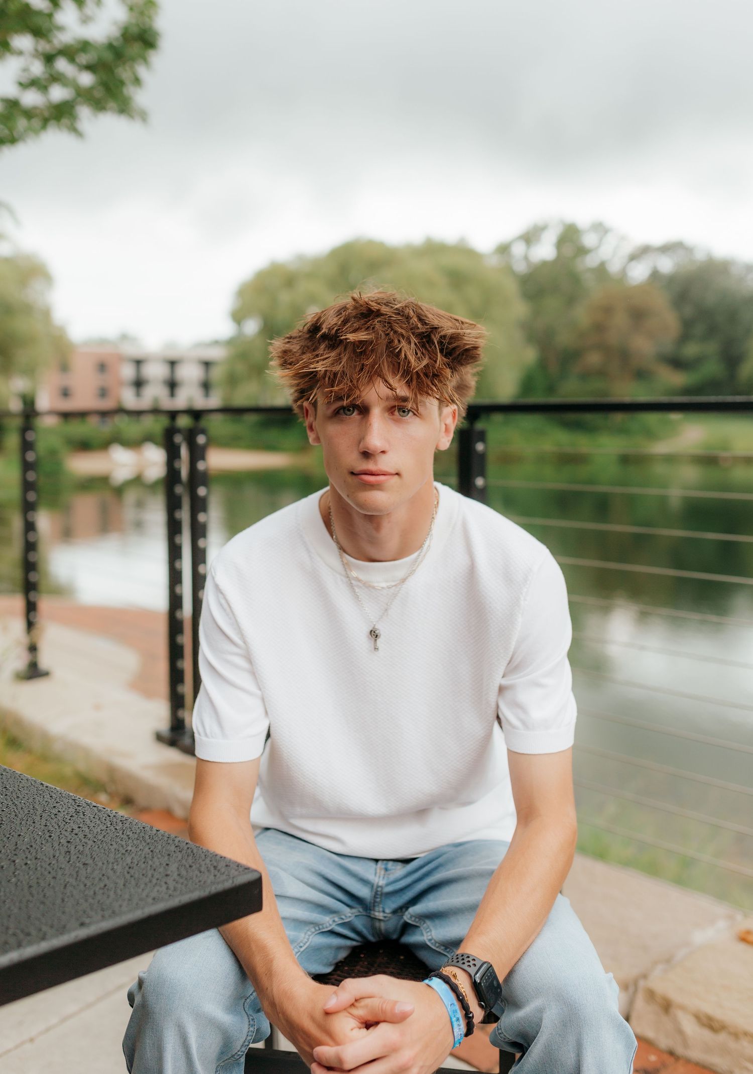 A young man with curly hair sits on a railing overlooking a body of water in an outdoor setting.