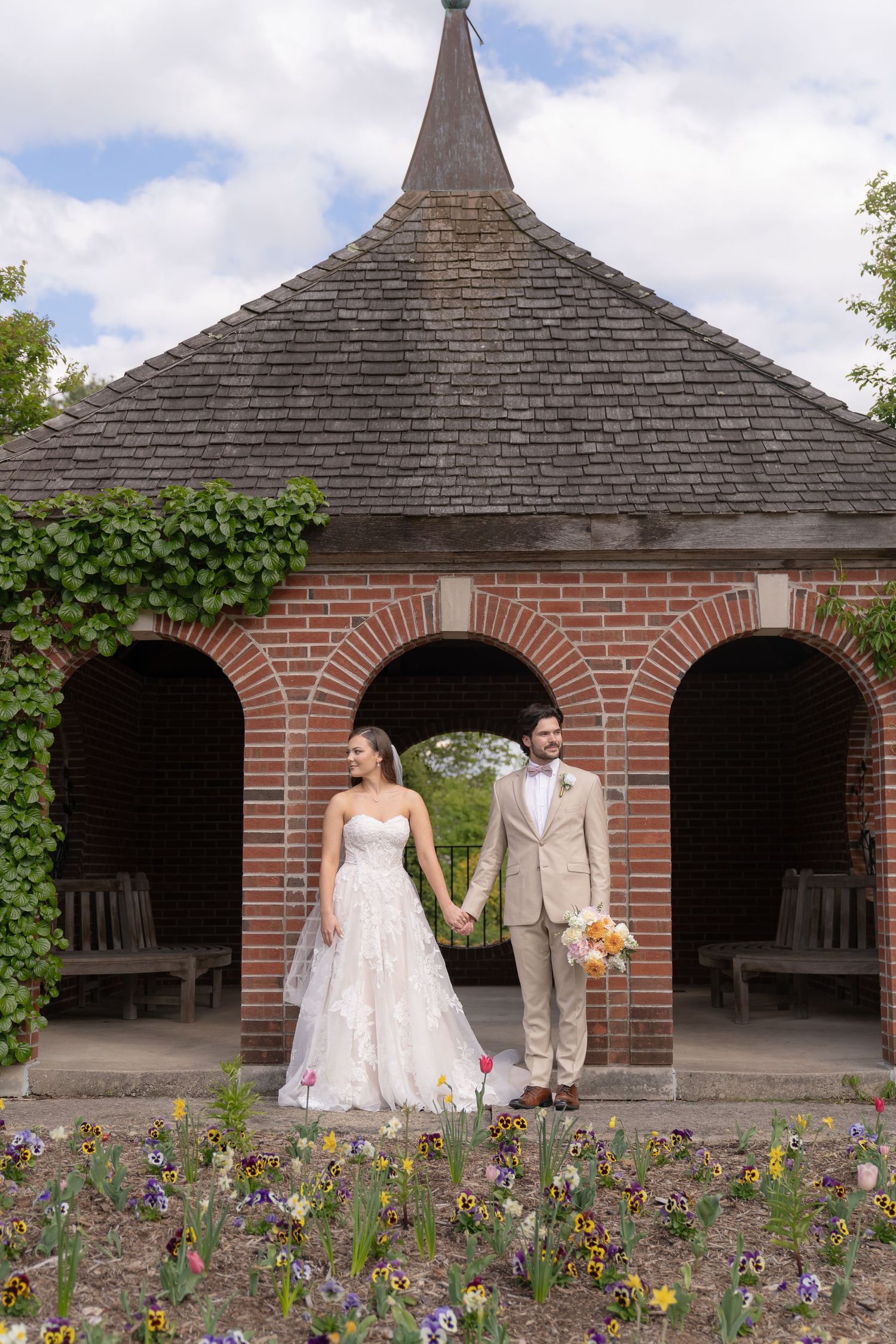 A bride and groom stand in front of a brick archway with a peaked roof in a garden setting.