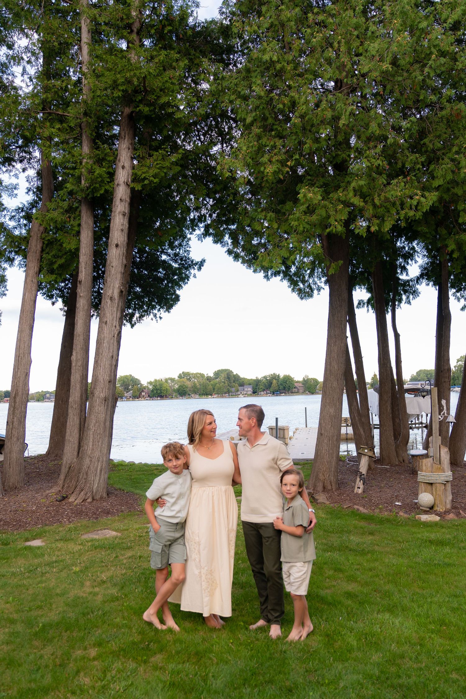 A family of four stands together on a grassy lawn surrounded by tall trees near a body of water.