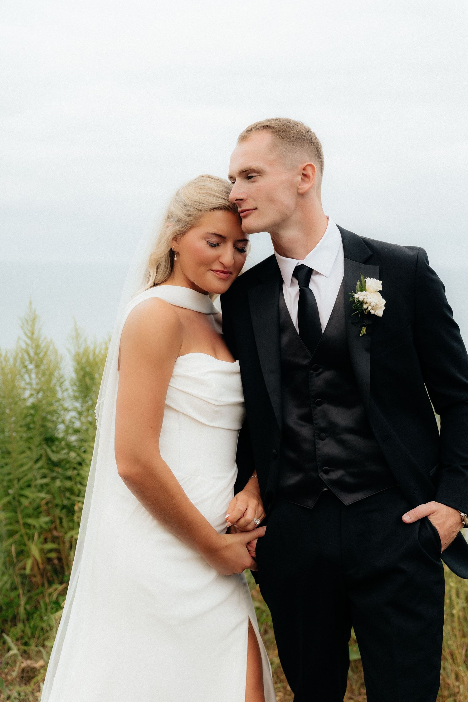 Bride in white gown and groom in black tuxedo embrace tenderly outdoors on their wedding day.