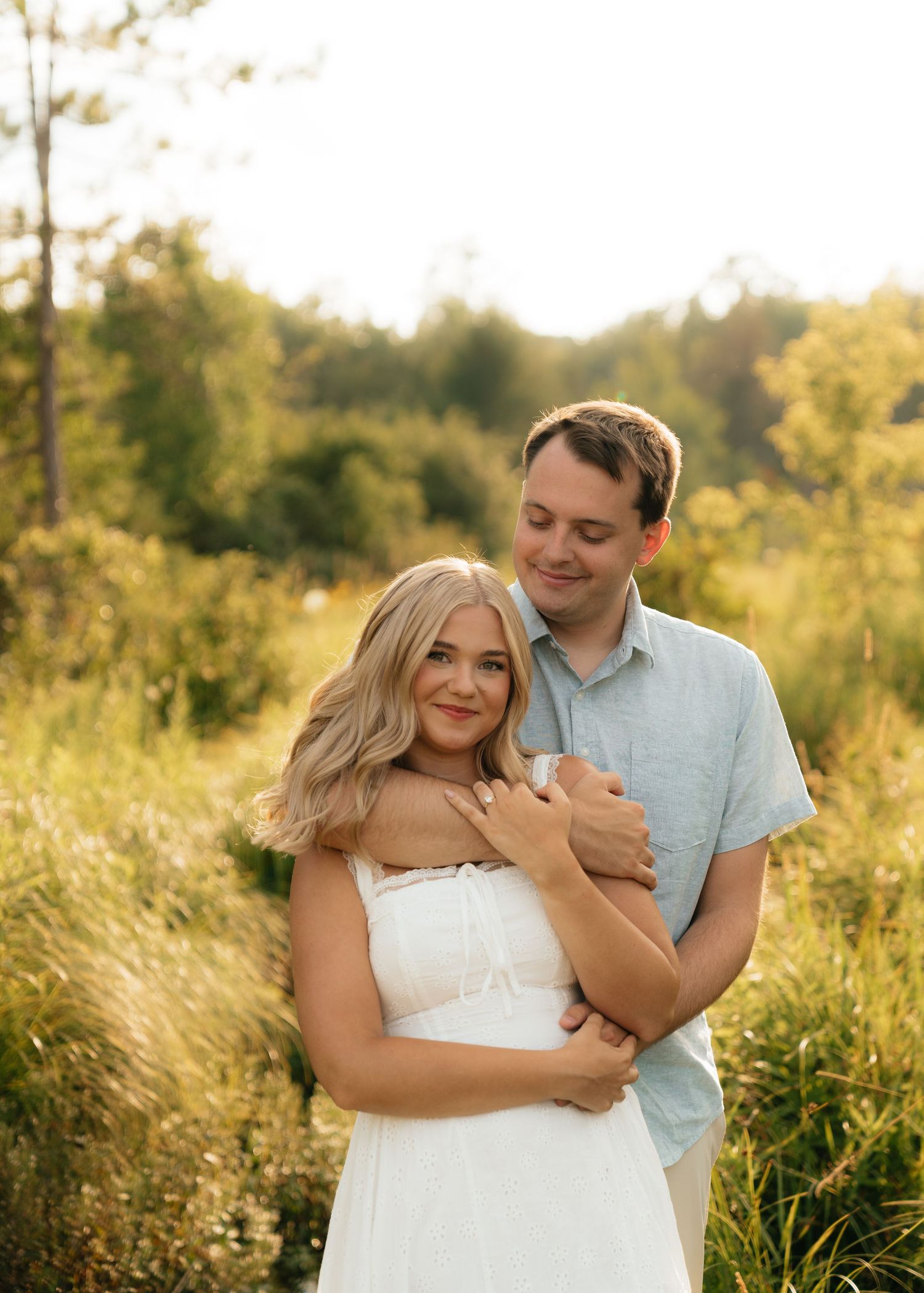 Couple embracing in a sunlit meadow, woman in white dress smiling, man in blue shirt, lush green background.