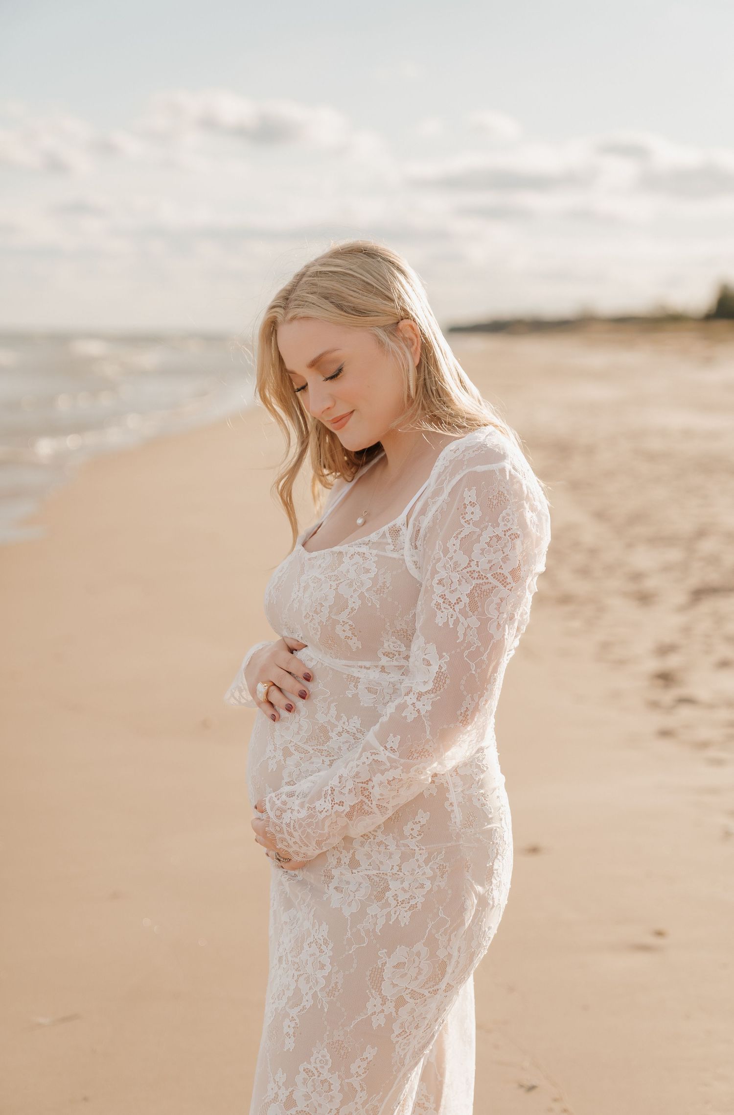Pregnant woman in white lace gown cradling baby bump on sandy beach at golden hour.