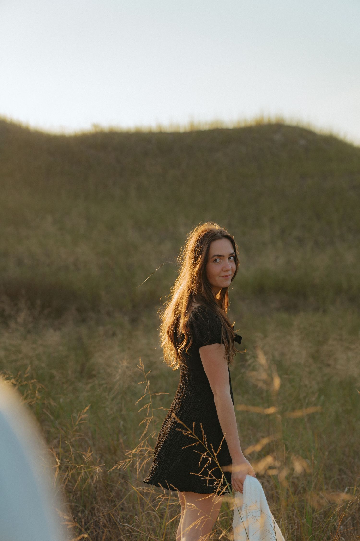 A woman with long brown hair stands in a grassy field, looking away from the camera with a thoughtful expression.