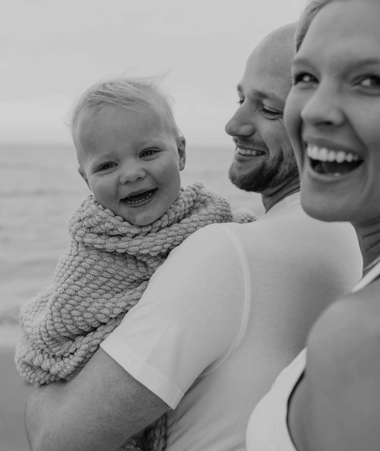 A happy family of three smiles and embraces on a beach, with a young child wrapped in a cozy knit scarf.