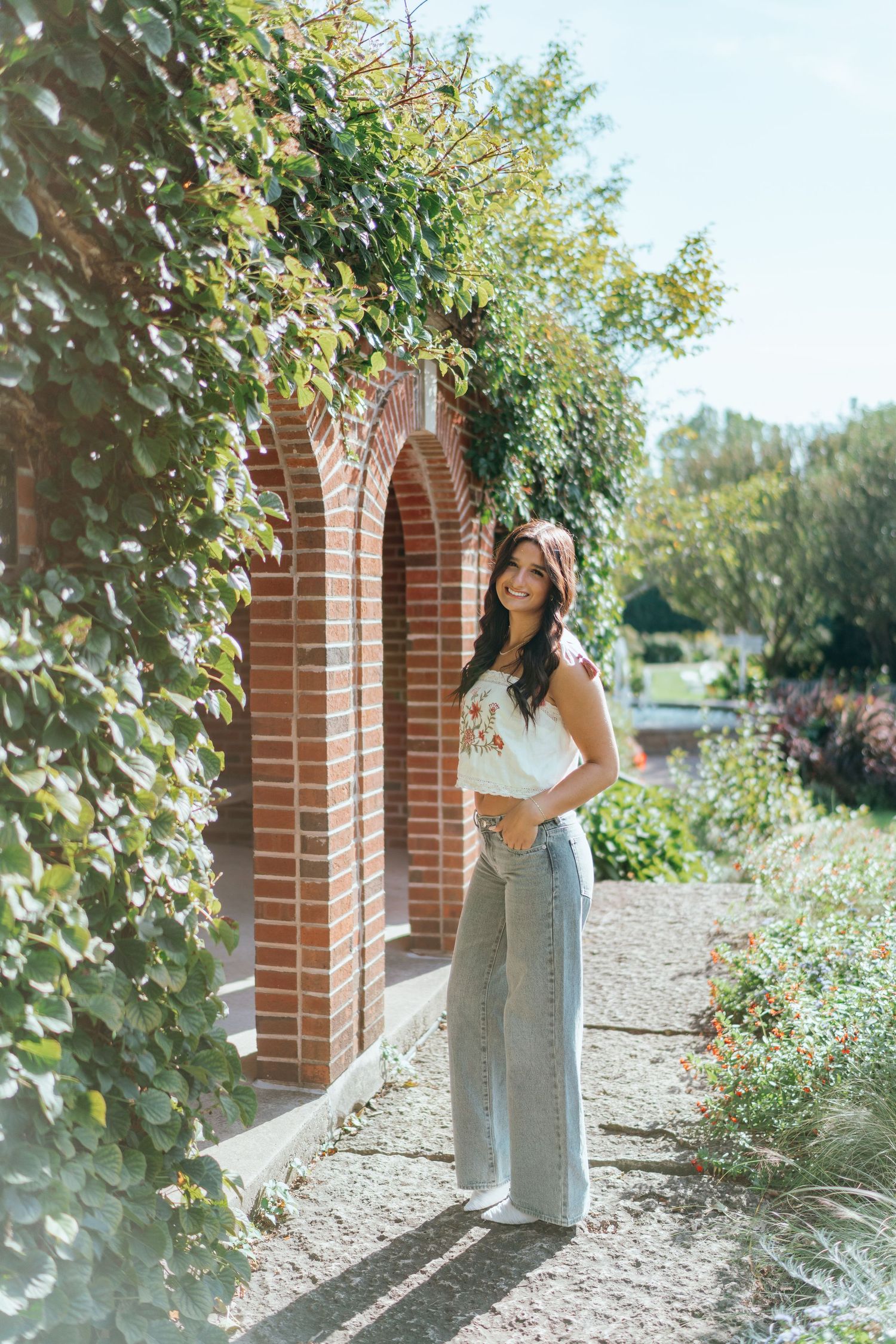 Young woman smiling in wide-leg jeans and floral top beside a ivy-covered brick archway in a sunny garden.