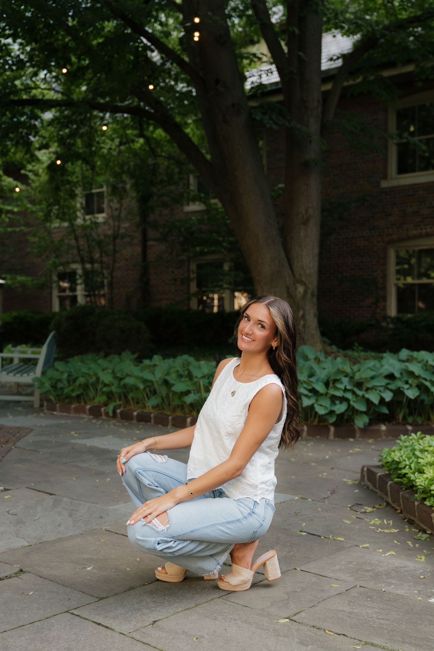 Young woman in white top and light jeans crouches smiling on stone patio near lush green trees and brick building.