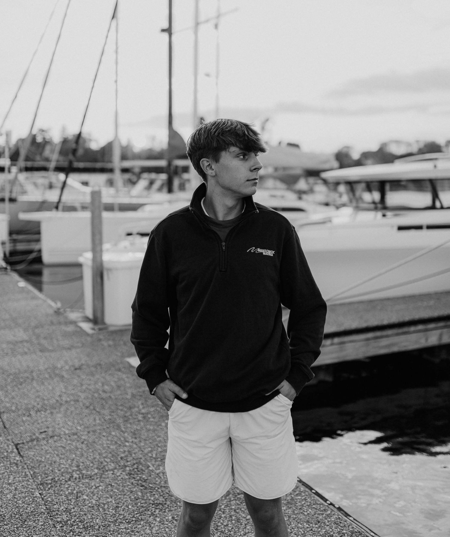 A young man wearing a black jacket stands on a dock near sailboats in a harbor.