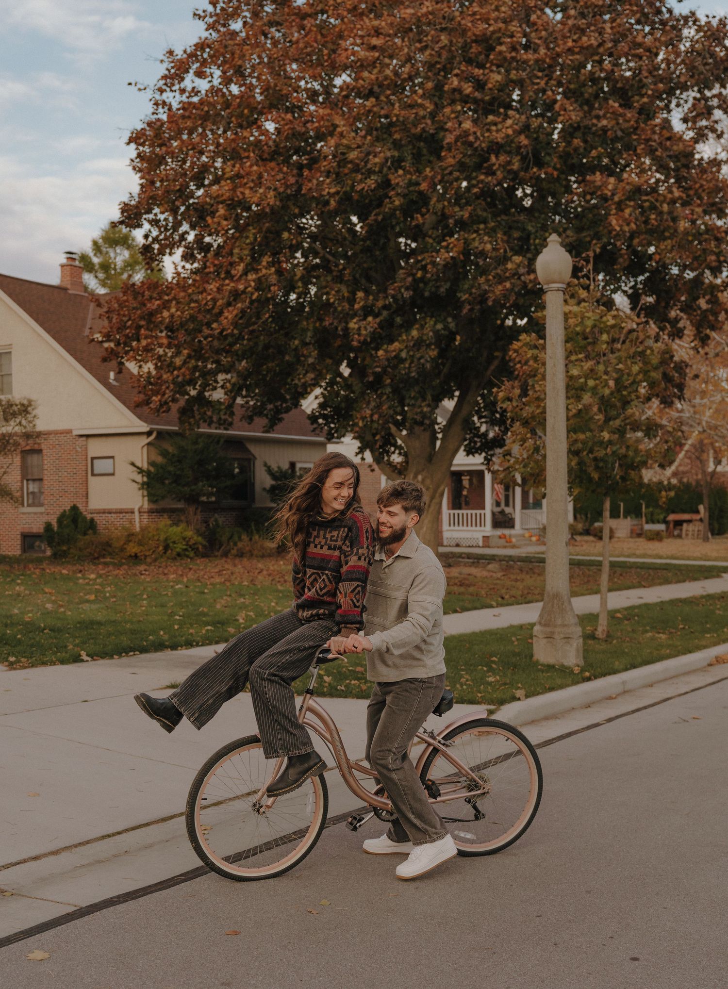 Two people riding a bicycle together on a paved path surrounded by trees and buildings.
