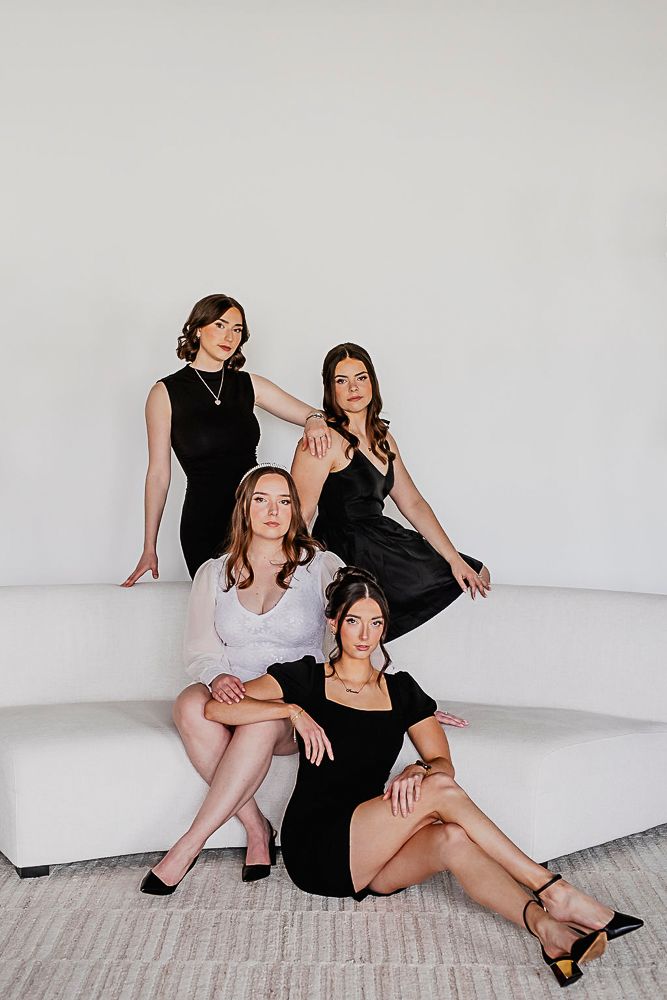Four women in elegant black and white dresses pose together on a white couch against a minimalist backdrop.