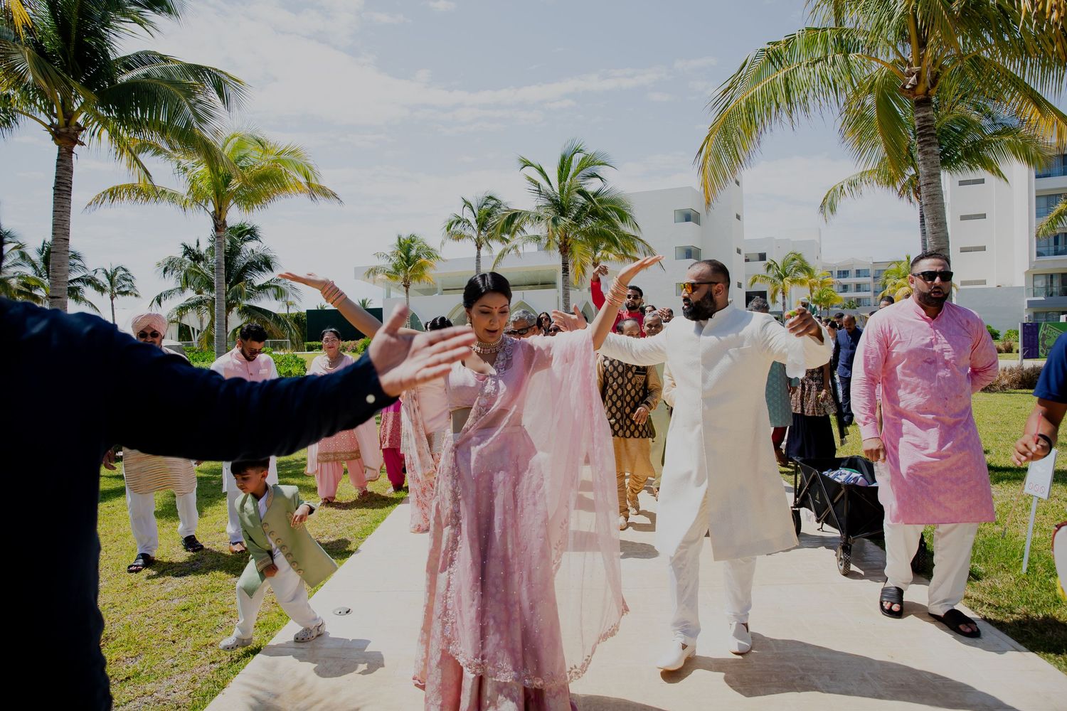 Festive outdoor wedding scene with guests in pink and white attire dancing among palm trees.
