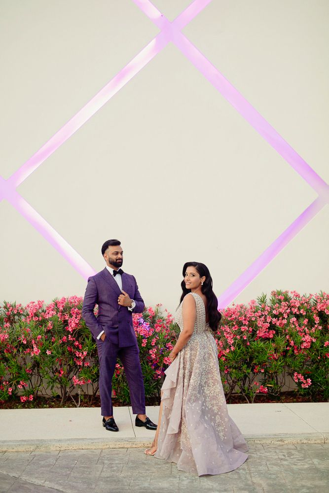 Couple in formal attire pose playfully against a geometric pink wall with red flowers.