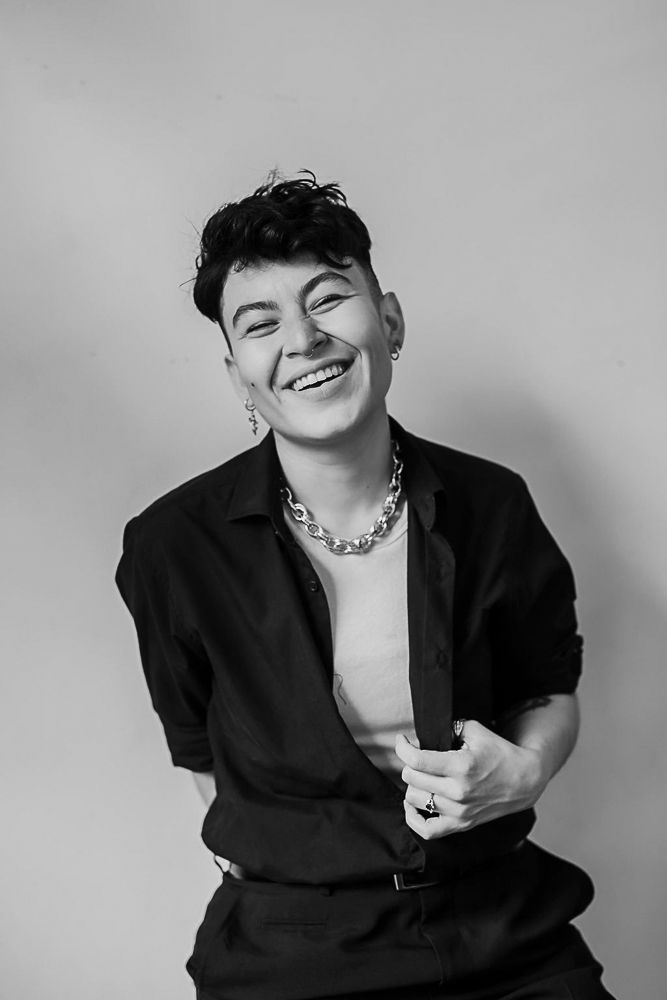Black and white portrait of person in dark shirt with chain necklace against plain background.