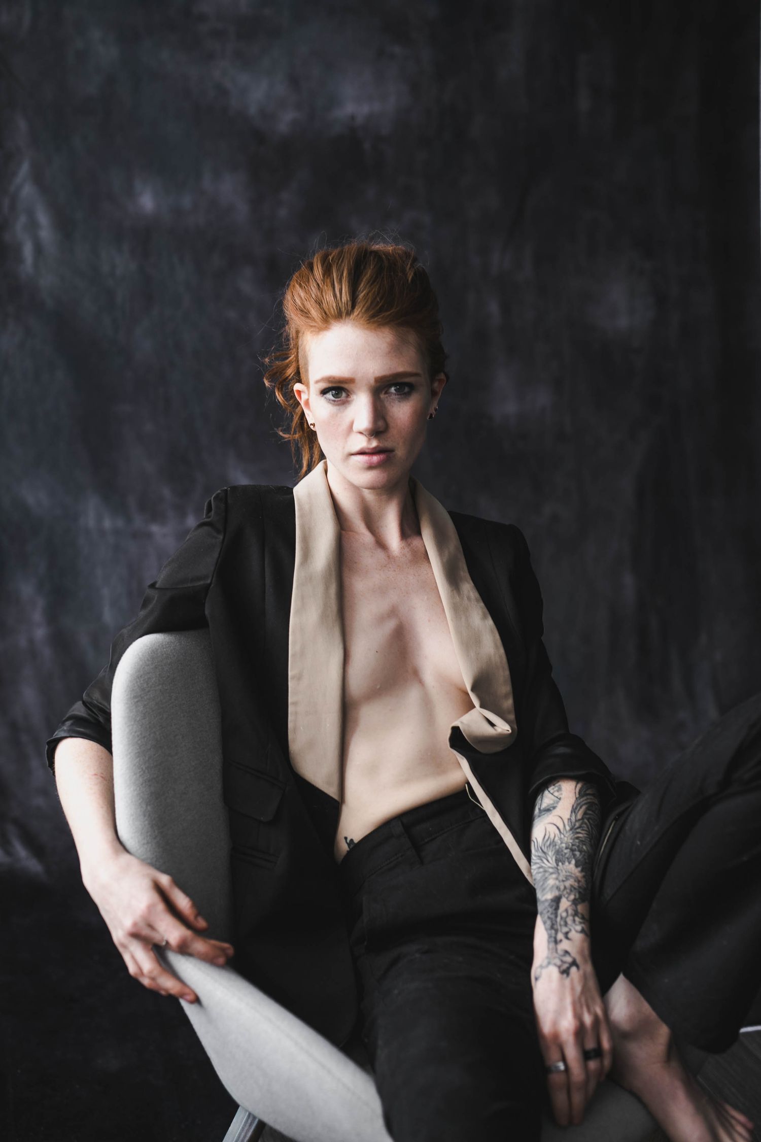 Redhead woman in black blazer with tattoo sits confidently on chair against dark studio background.