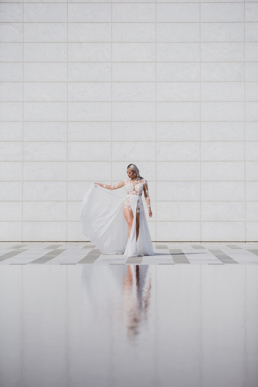A figure in a flowing white dress stands against a minimalist concrete wall, creating an ethereal reflection on the floor.